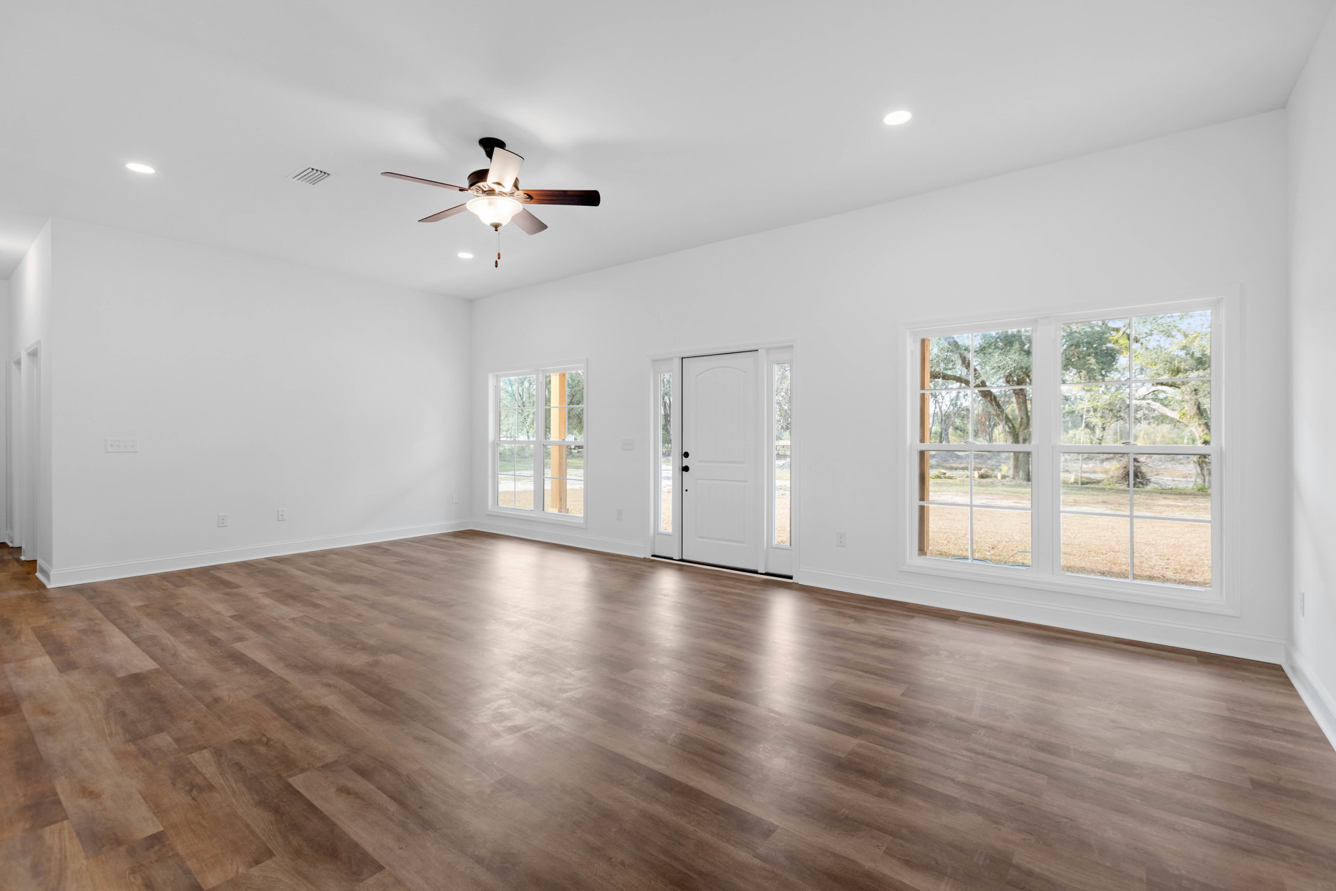 Ceiling fan with light fixture above wood laminate flooring, white door with black knobs, large windows showing trees outside