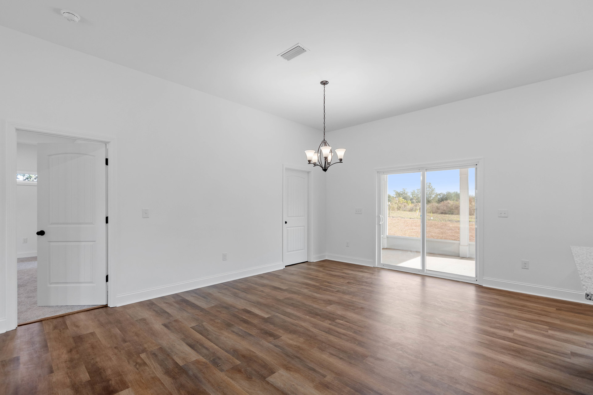 Wood flooring in a bright room with a modern chandelier, white walls, a white door with black hardware, ceiling vent, and sliding glass door.
