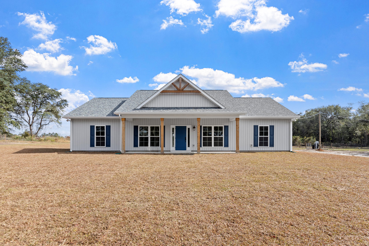 Two-story house with triangular roof, blue front door and white trim, blue shuttered windows, surrounded by large grassy yard and mature trees under a partly cloudy sky