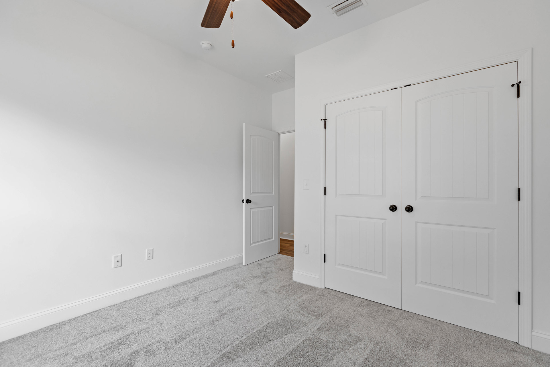 White-walled room with ceiling fan, open white double doors with black knobs, carpeted floor, and wooden curtain against white background