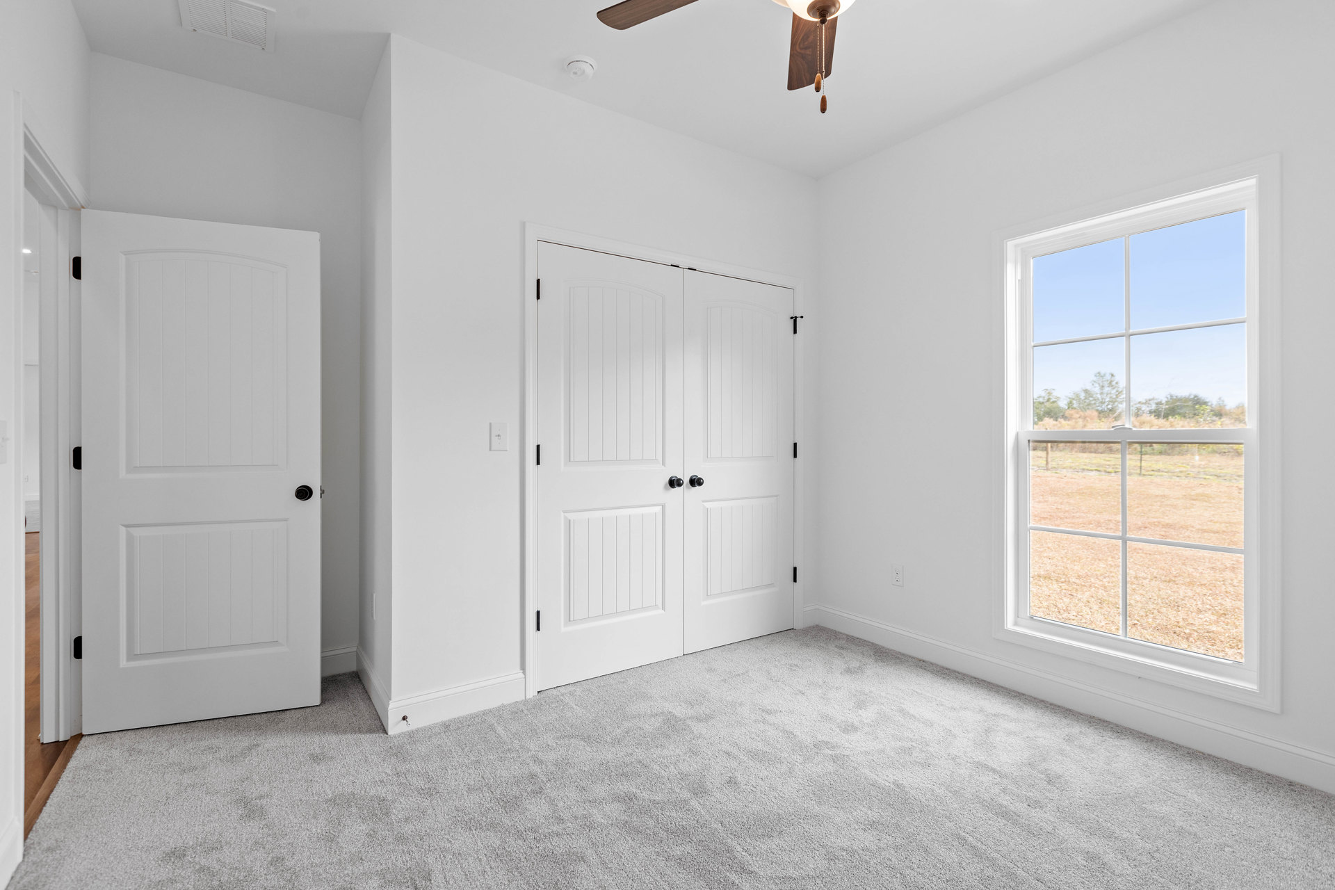 Bright room with white carpet, ceiling fan, single white door with black handle, double white doors with black knobs, and window overlooking grassy field