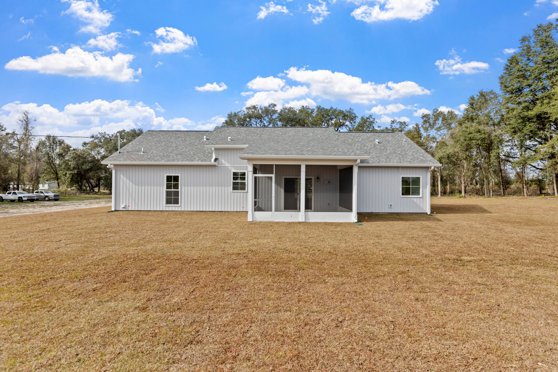 Two-story house with gray siding, screened porch, large grassy yard, mature trees in background, white and silver trucks parked on lawn, close-up window visible.