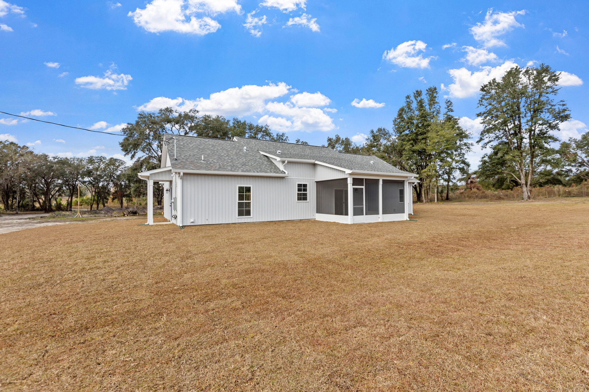 White siding house with screen door, expansive grassy yard, mature tree with spreading branches, cloudy sky, roof partially visible with trees in background