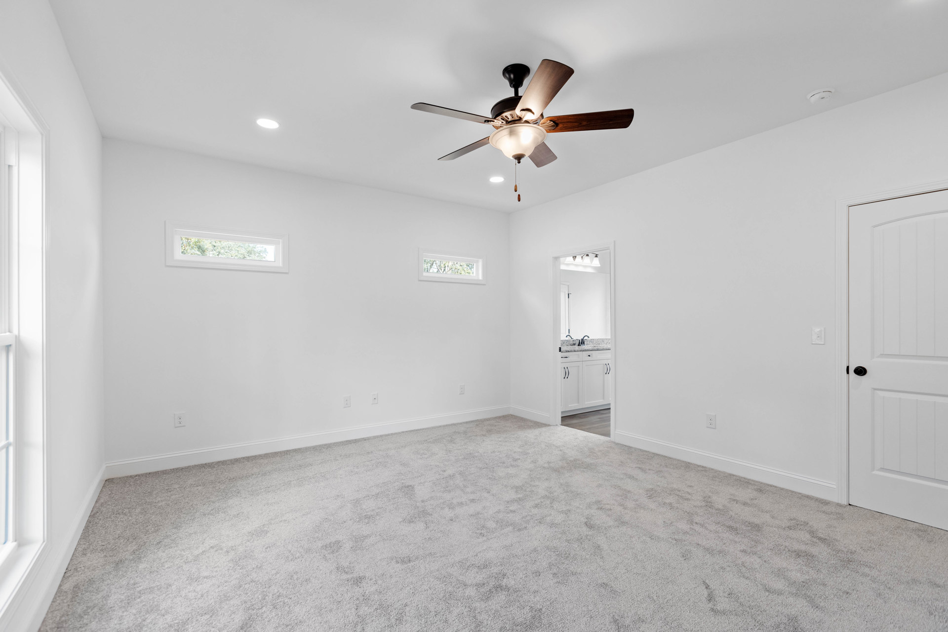 Carpeted bedroom with white walls, ceiling fan and light fixture, large window overlooking trees, and black lamp shade