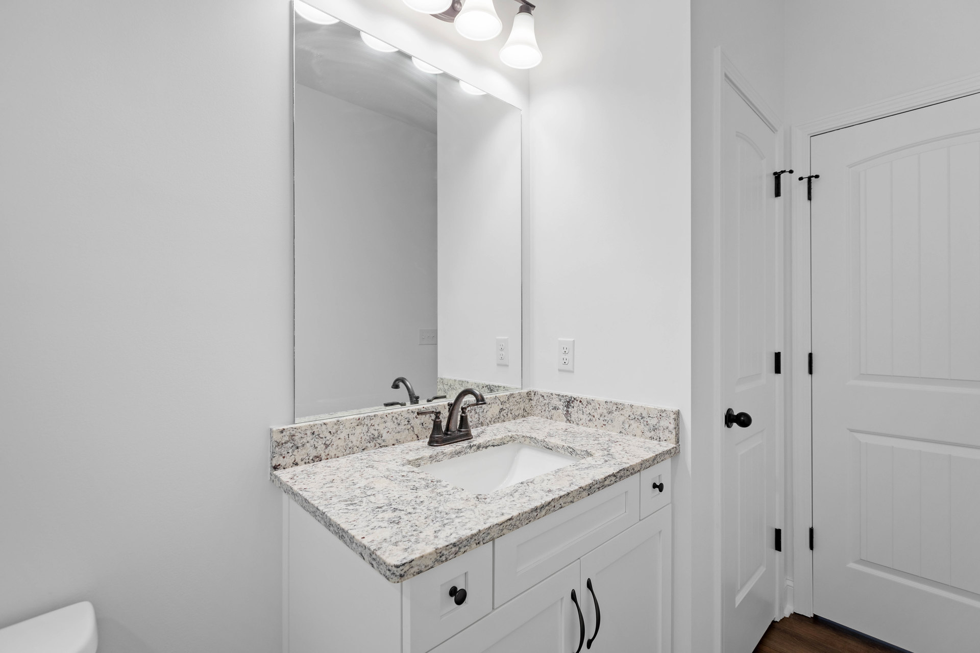 Marble countertop with undermount white sink, black faucet and hardware, large framed mirror above, white cabinetry, tile backsplash, ceiling light fixture, white door with black
