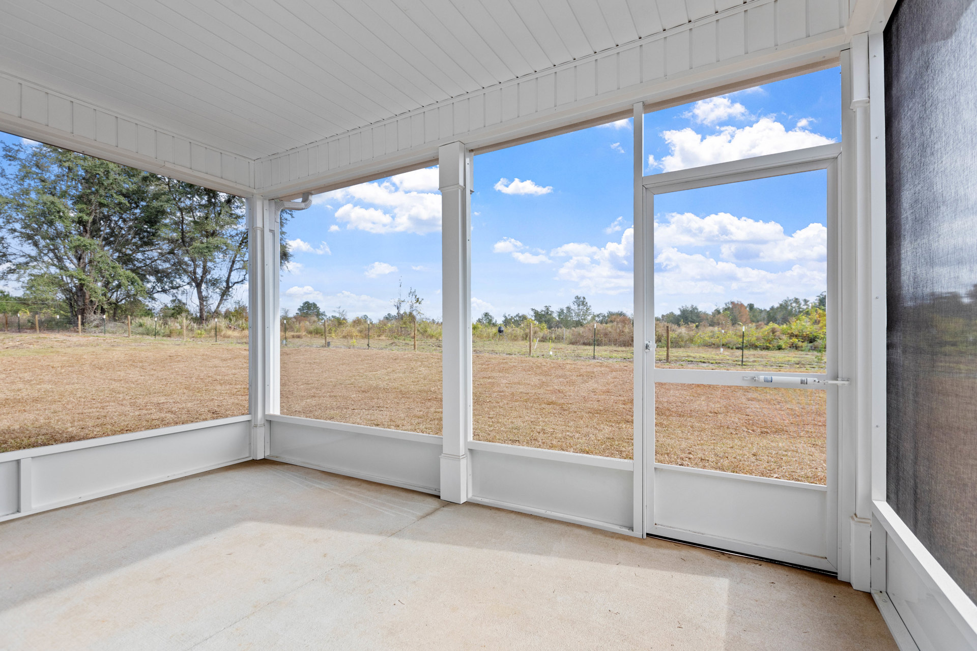 Spacious room featuring large floor-to-ceiling windows overlooking a dirt field, white pillar near a grill, fence and trees visible outside, light-colored flooring and composite