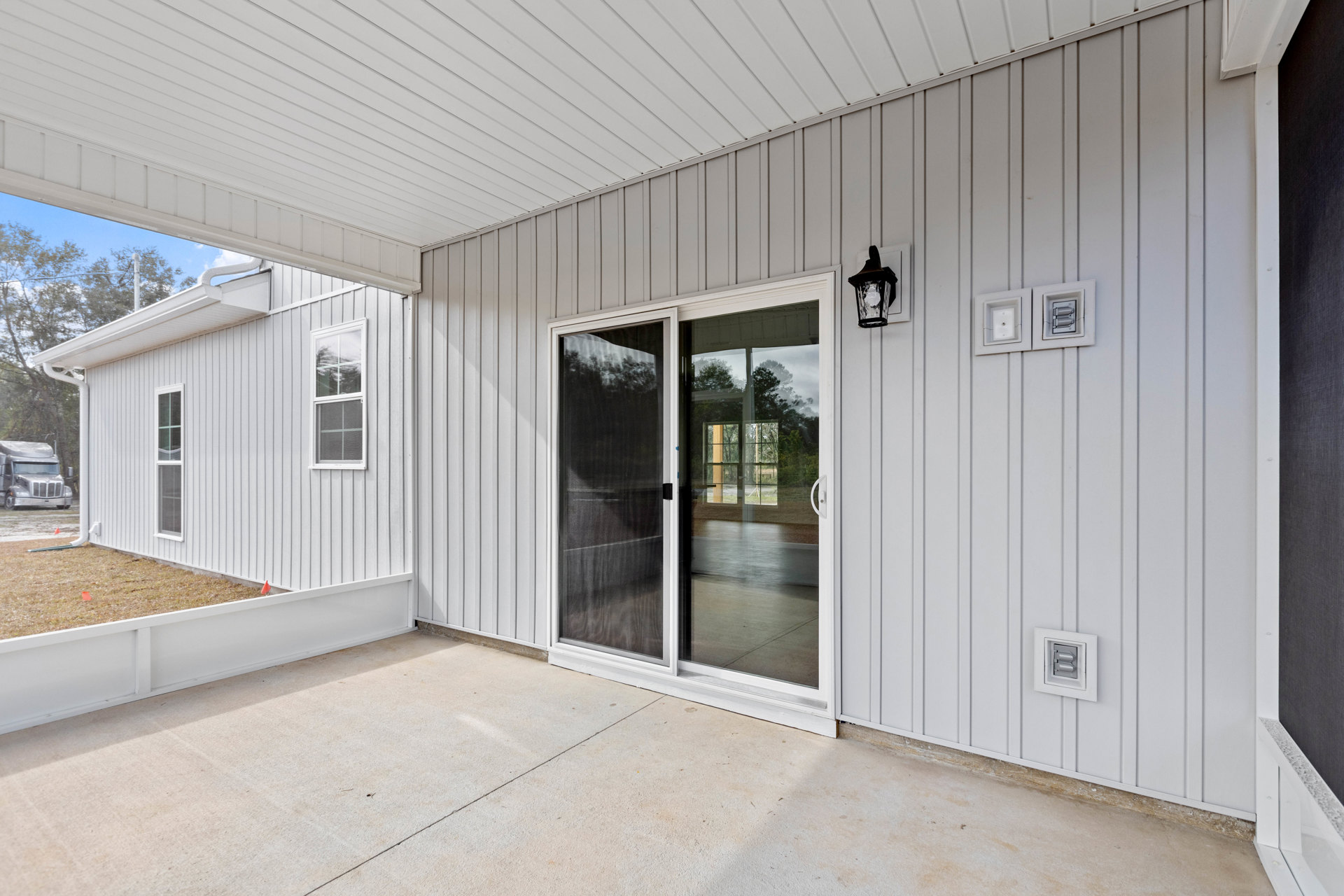 White exterior with smooth siding, sliding glass door with screen, square wall fixture, adjacent window, and ceiling light visible.
