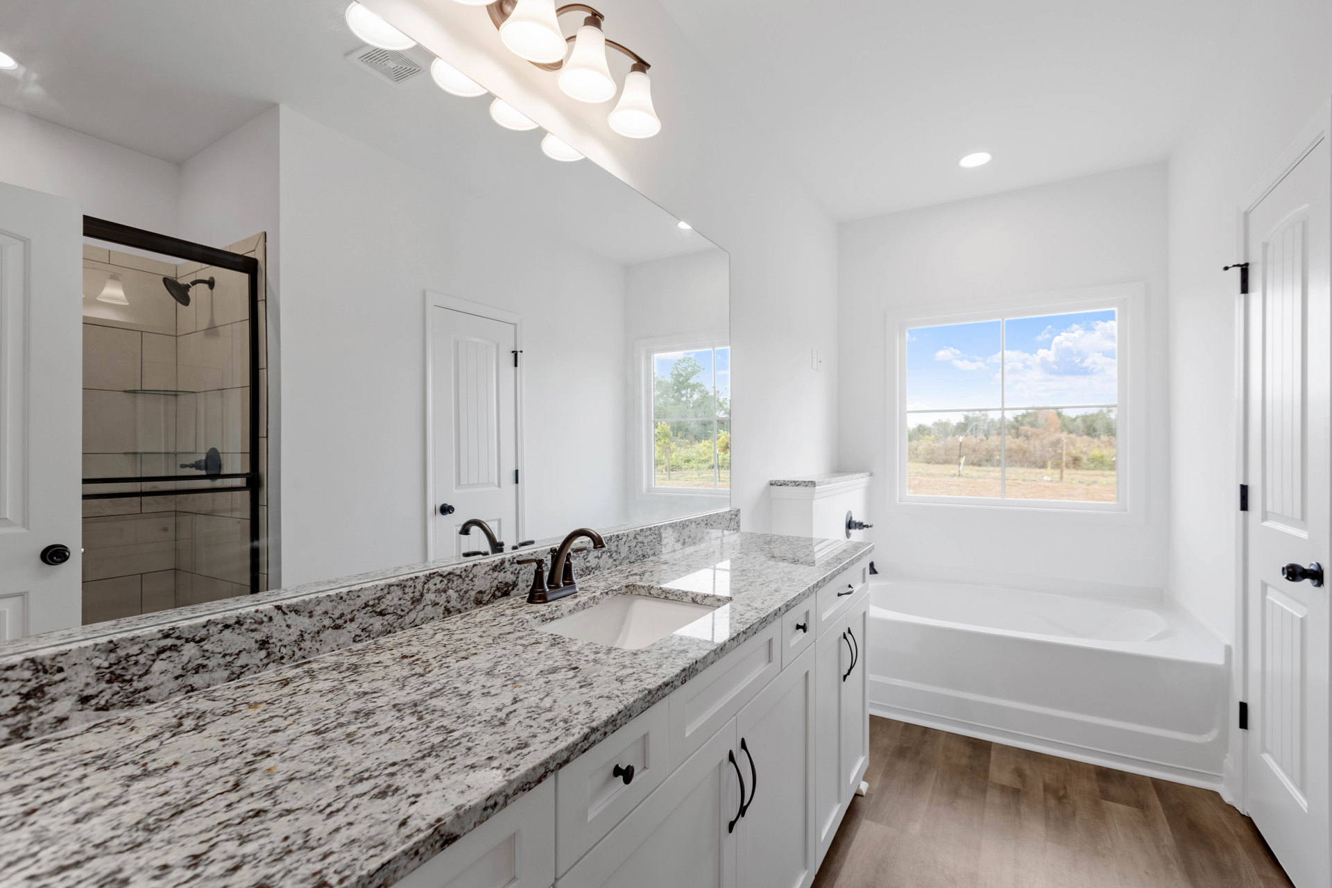 Bathroom with marble countertop, white sink, glass-door shower, freestanding tub, and window overlooking field and trees, illuminated by modern light fixture