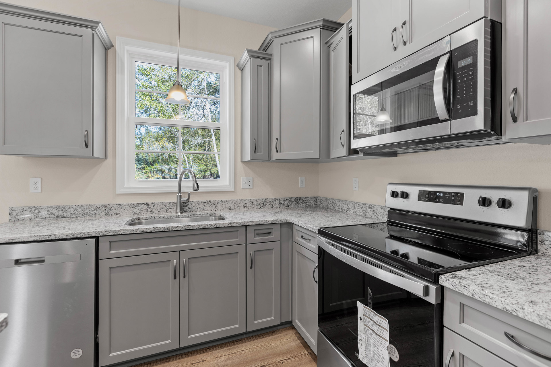 Grey kitchen cabinets with granite countertops, stainless steel microwave above cabinetry, stove and oven, ceiling light fixture, white door with black handle, sink set in kitchen
