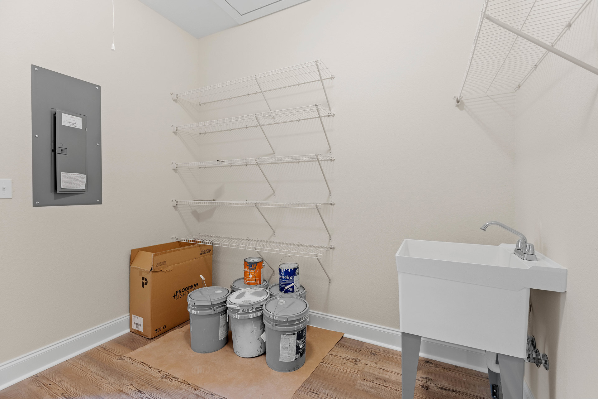 White-walled room with built-in white shelves, grey door displaying a white sign, large cardboard box beside a bucket, paint cans and roller on the tile floor, grey bucket with