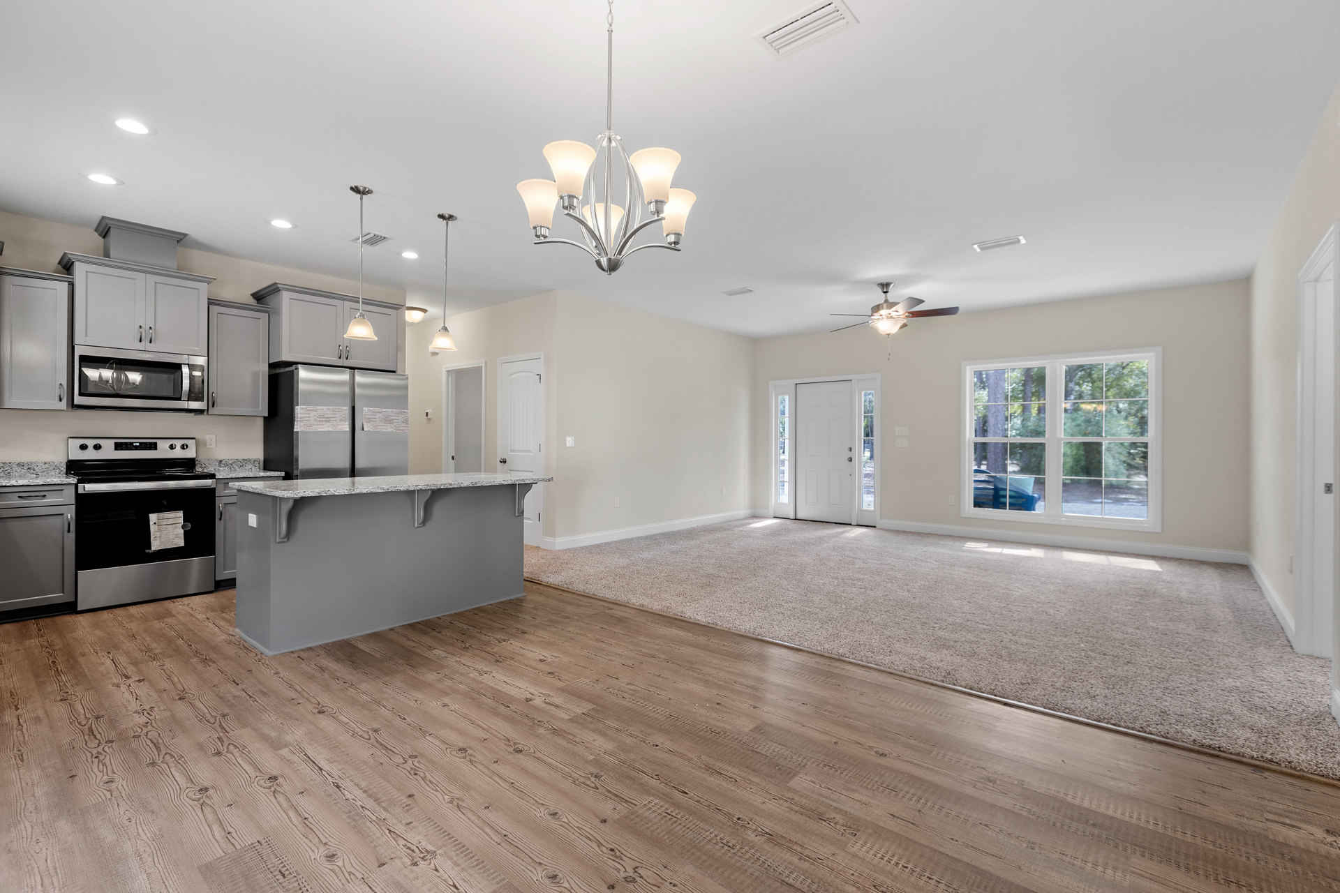 Open concept living room and kitchen with wood flooring, white cabinetry, stone countertops, stainless steel stove, five-light ceiling fixture, and large window with outdoor bench