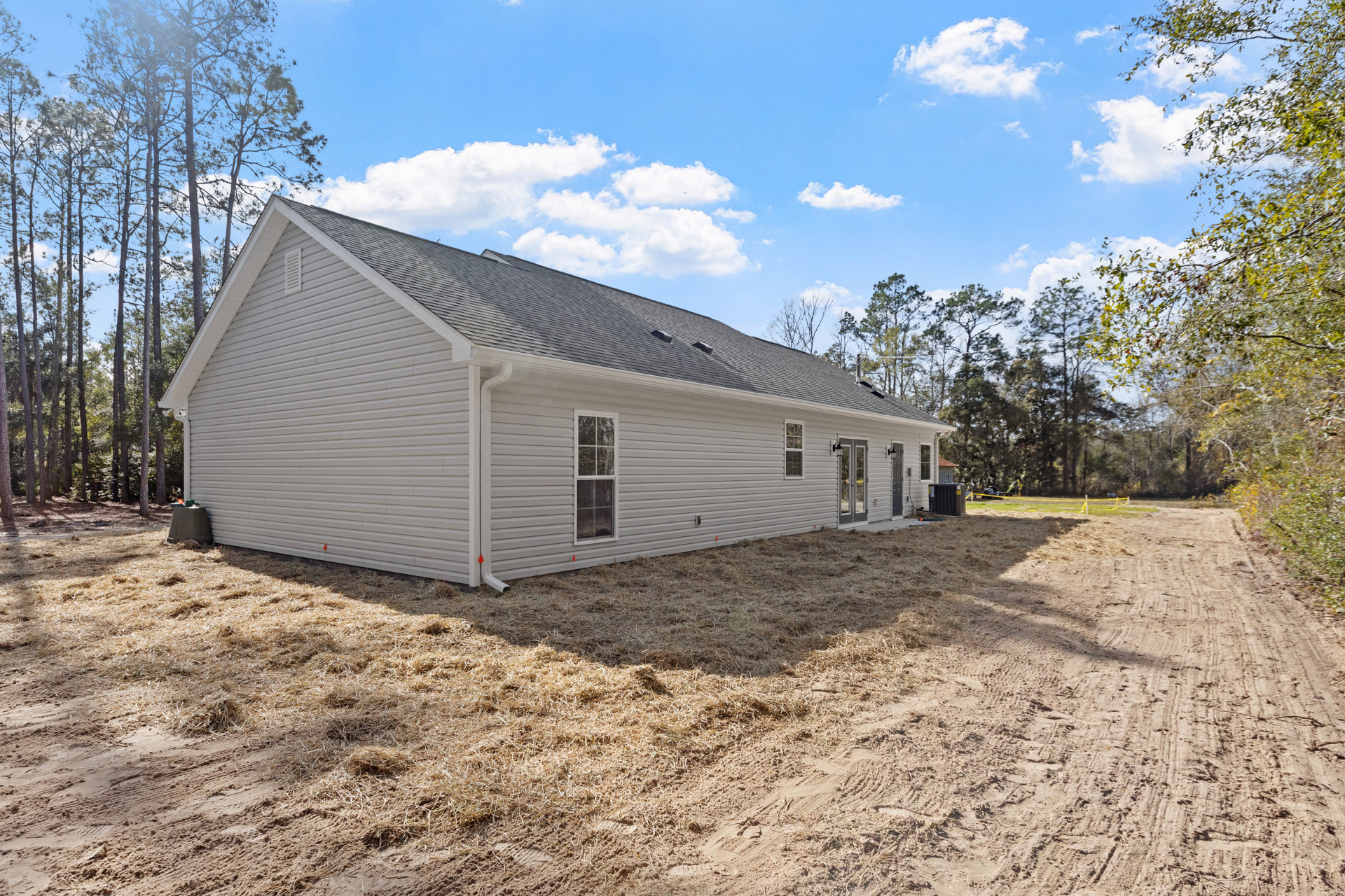 Partially built house with exposed framing, white window trim, dirt and grass yard, roof installed, trees and blue sky with clouds in background, green-topped trash can near