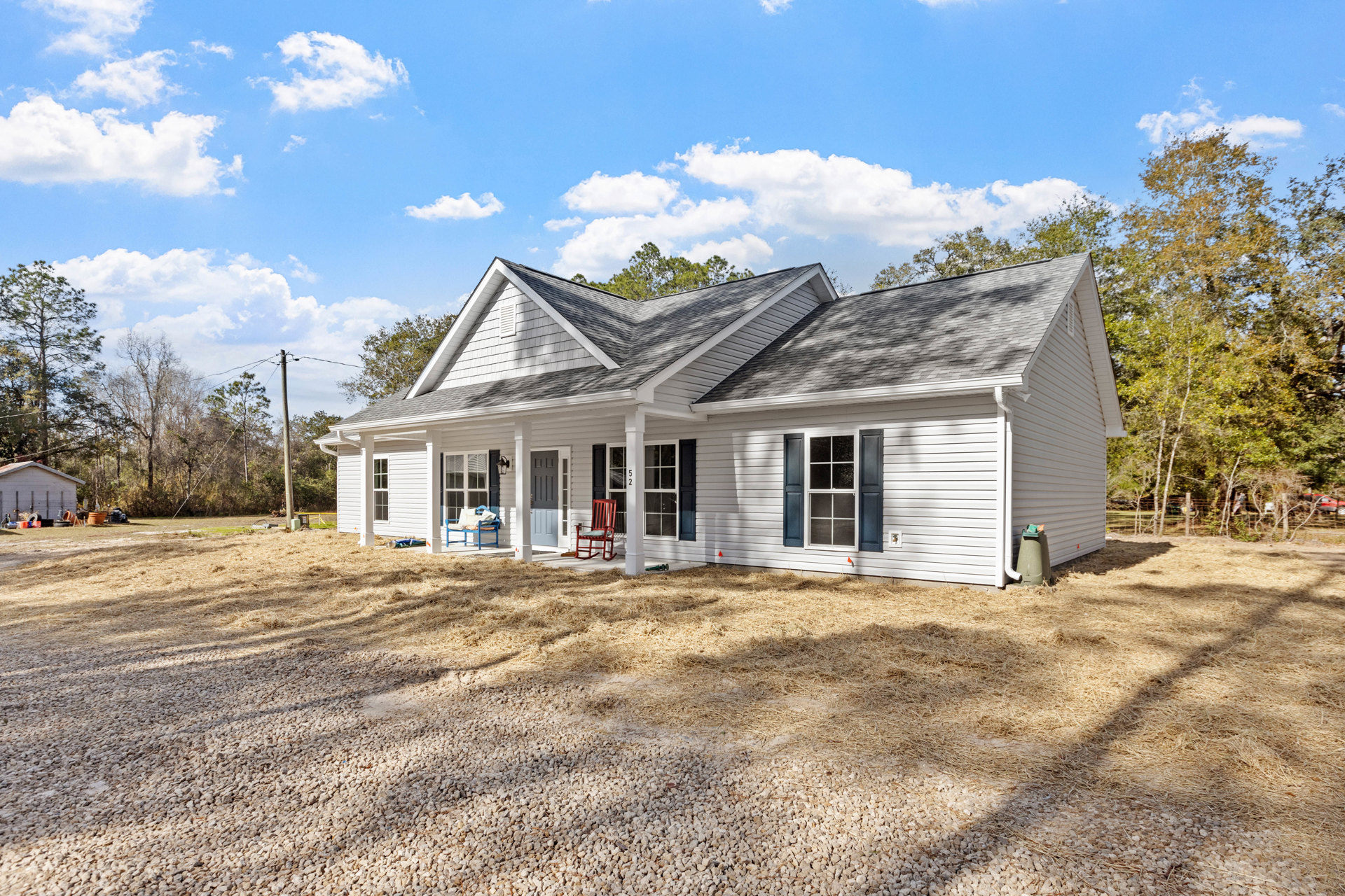 White cottage-style house with black shutters, white front door, red rocking chair on covered porch, gravel driveway, and trees in the background.