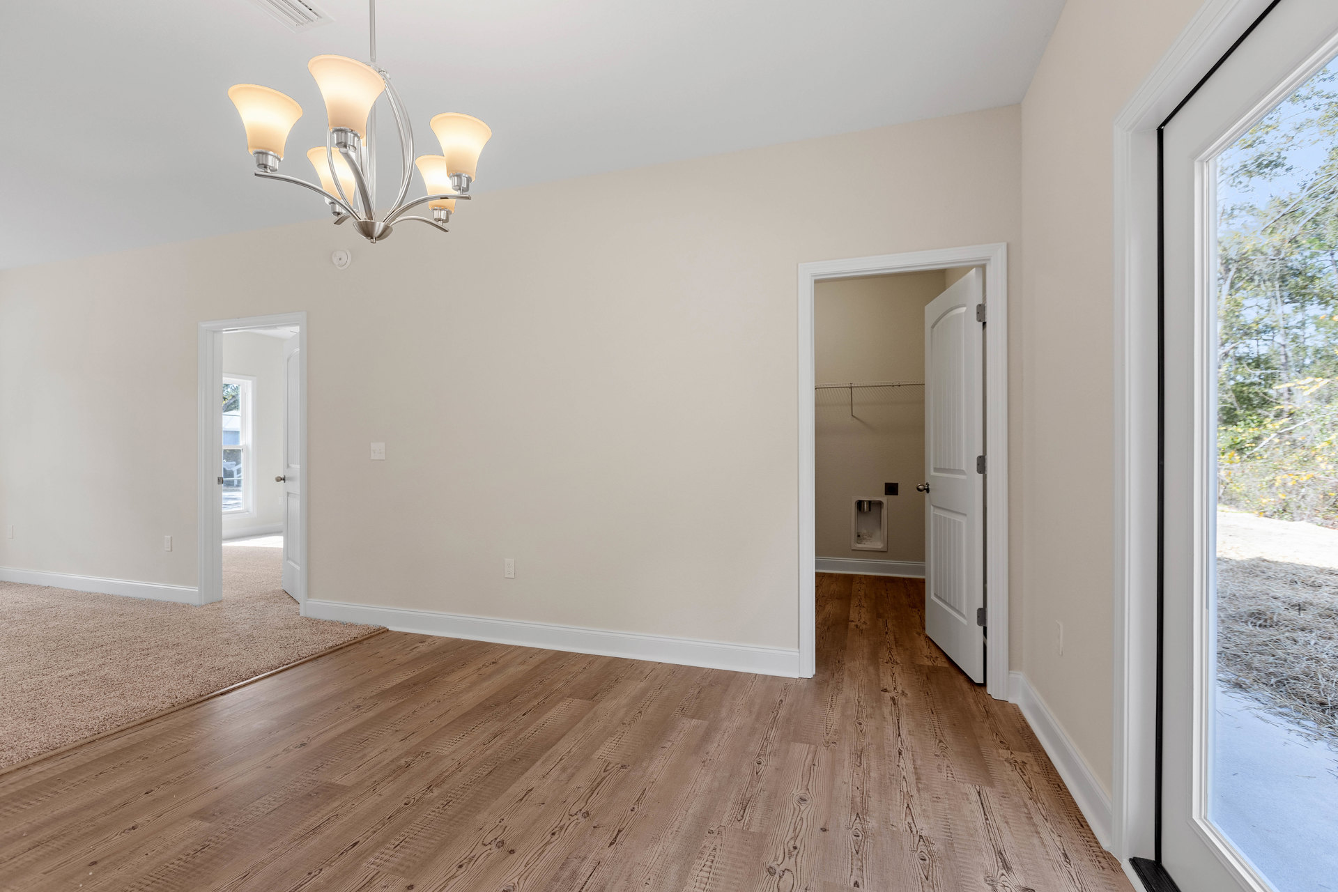 Chandelier with white shade hanging in a room featuring wood flooring, white plaster walls, a door with a doorknob, and a window overlooking trees.