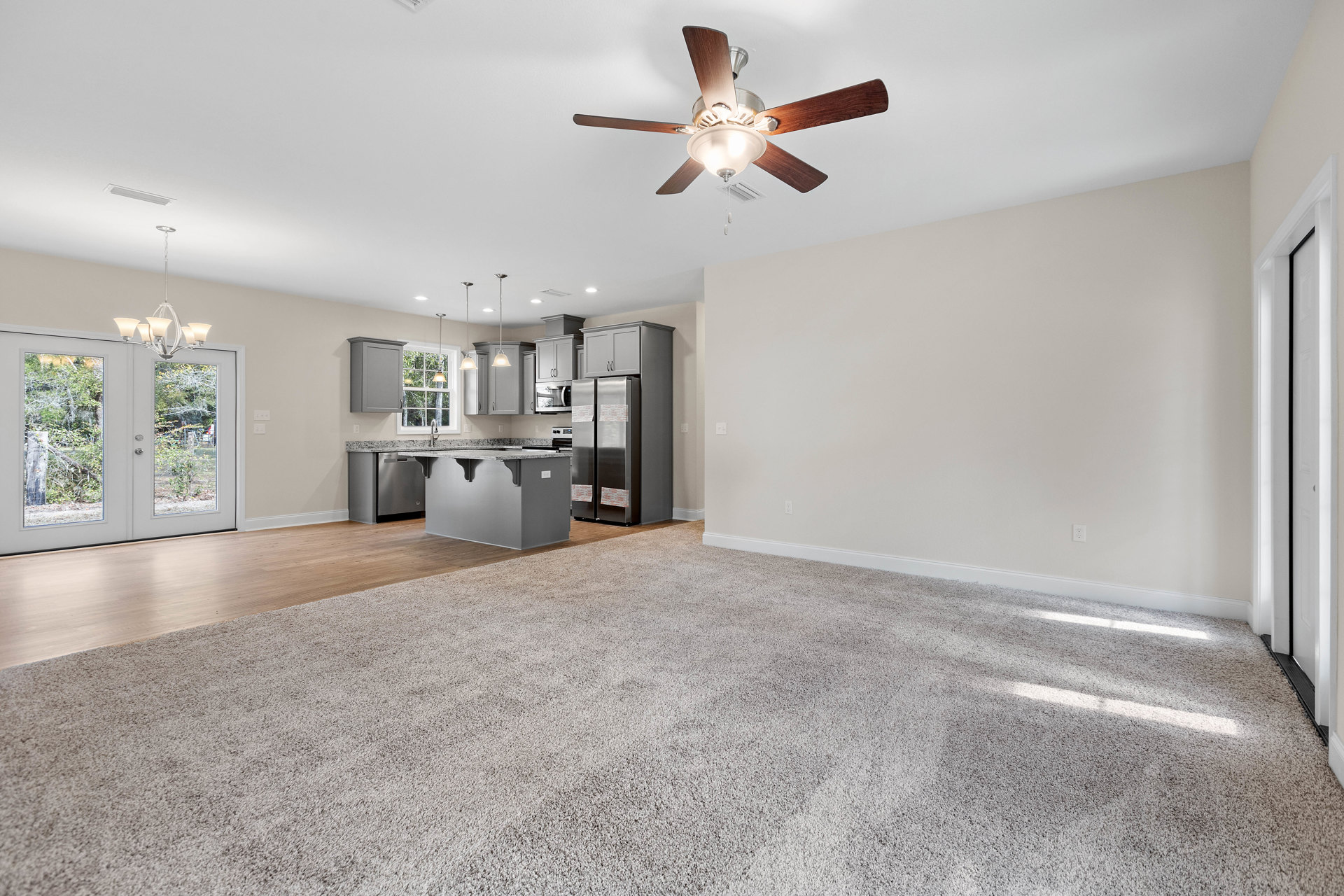 Living room with carpet flooring, ceiling fan with light fixture, open kitchen featuring stainless steel refrigerator, gray island with marble countertop, and double glass doors.