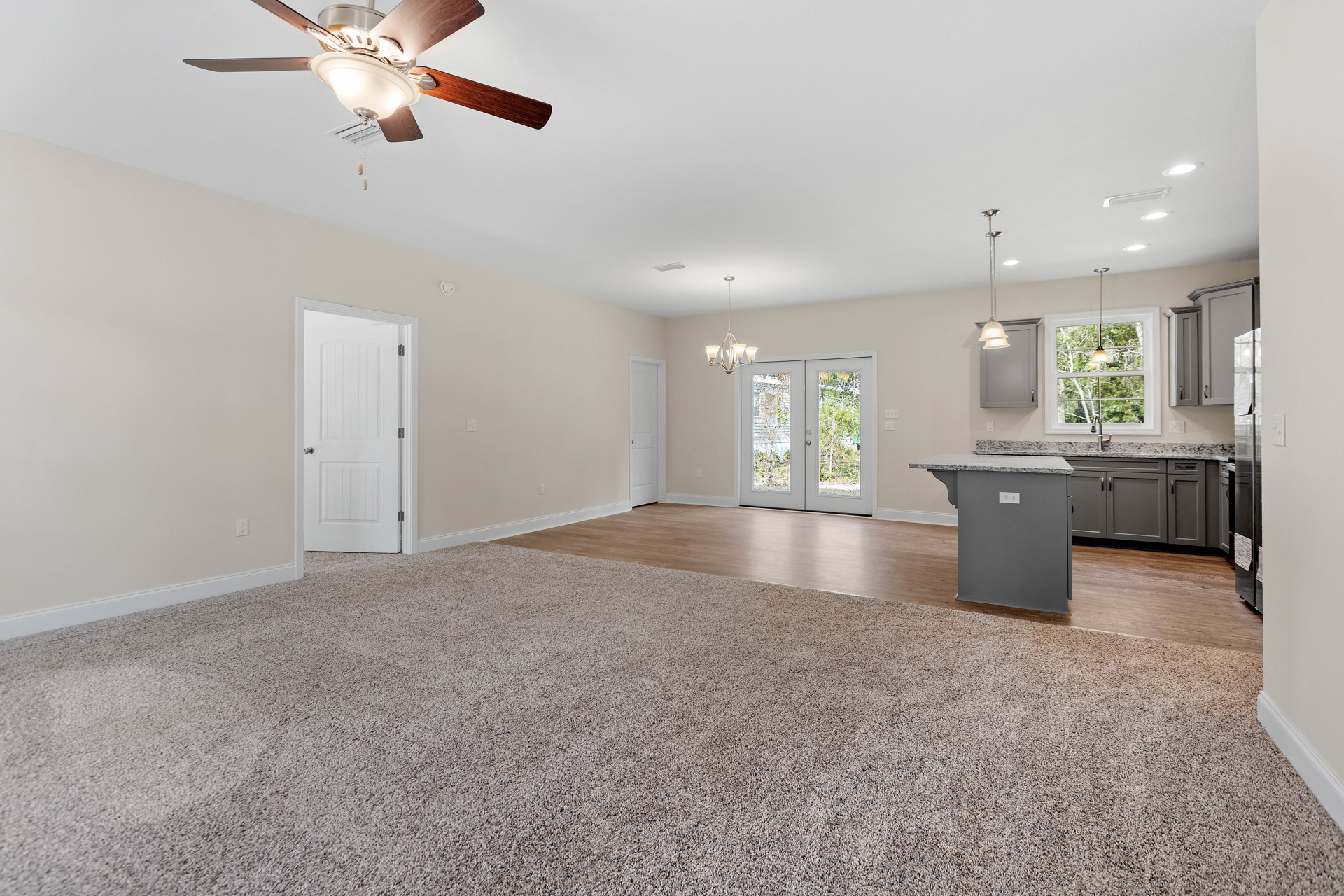 Open concept living room with carpet flooring, adjacent kitchen featuring a marble-topped island, white cabinetry, double glass-paneled doors, and ceiling fan with light fixture