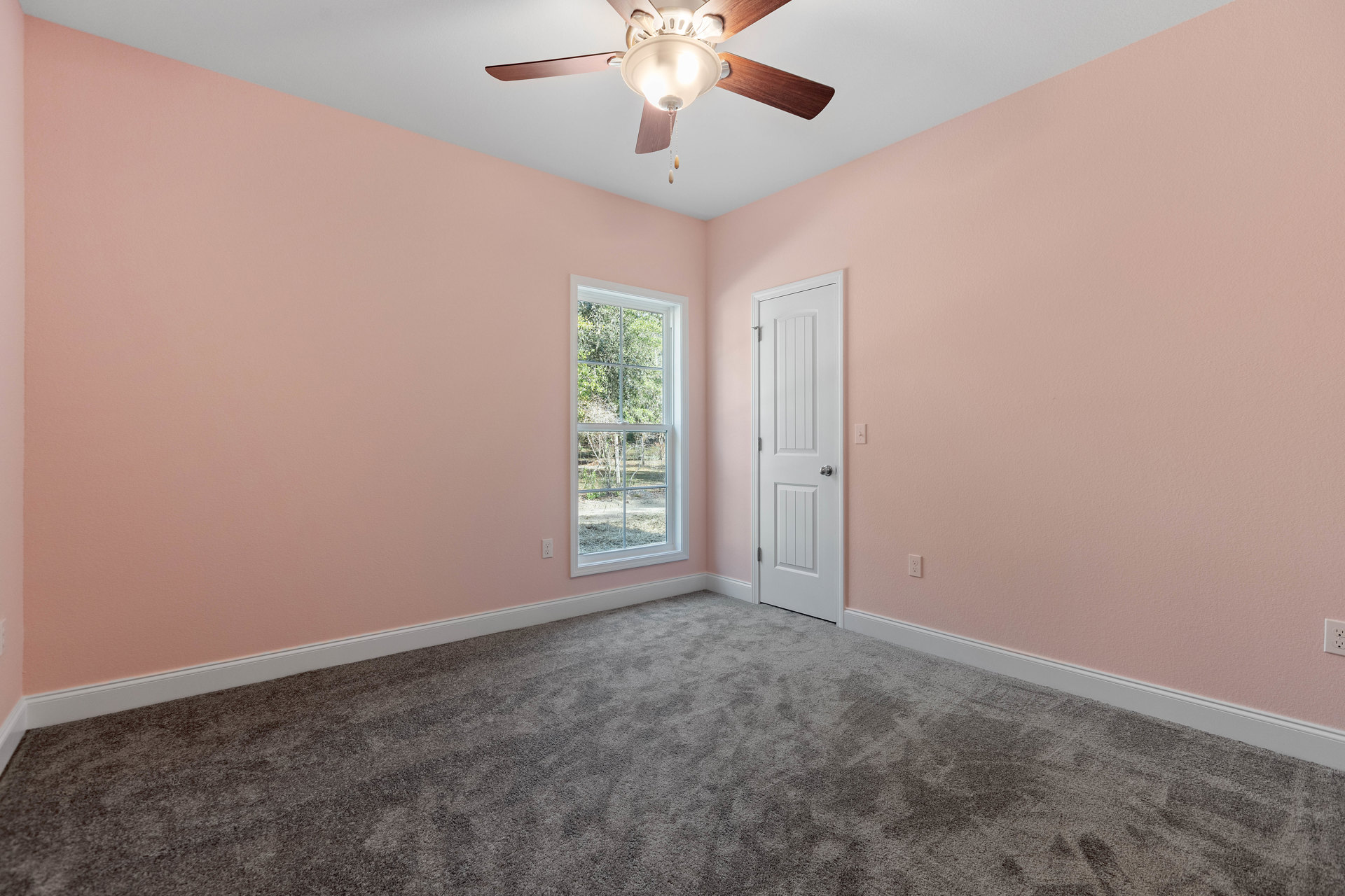 Bedroom with pink walls, white trim, carpeted floor, ceiling fan with light fixture, window overlooking trees, and white door with silver knob