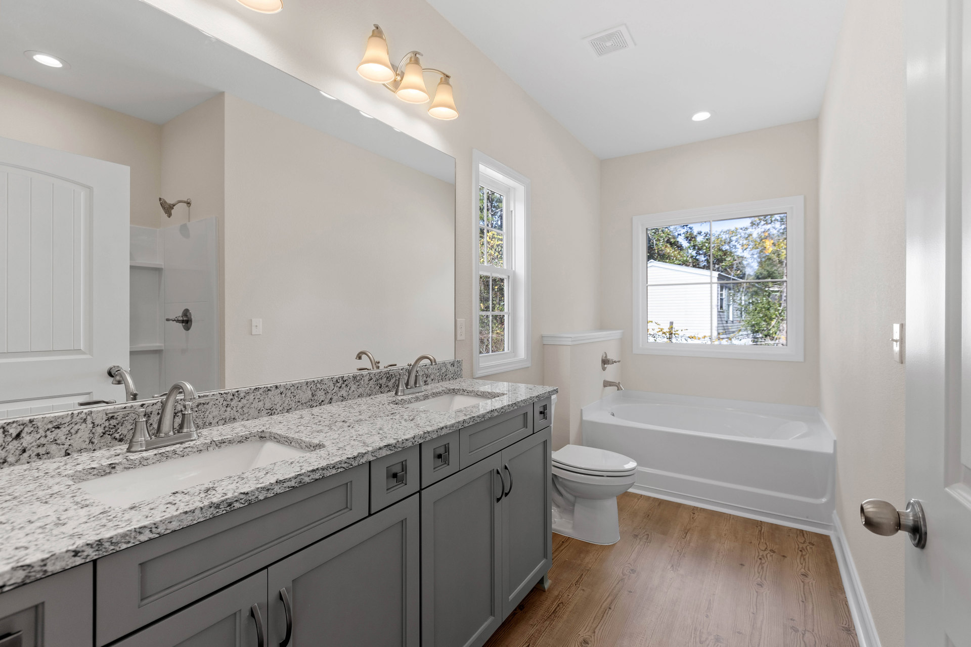 Bathroom with white bathtub and toilet, rectangular window framed in white showing trees outside, three-light fixture above sink, light-colored tile walls and countertop, modern