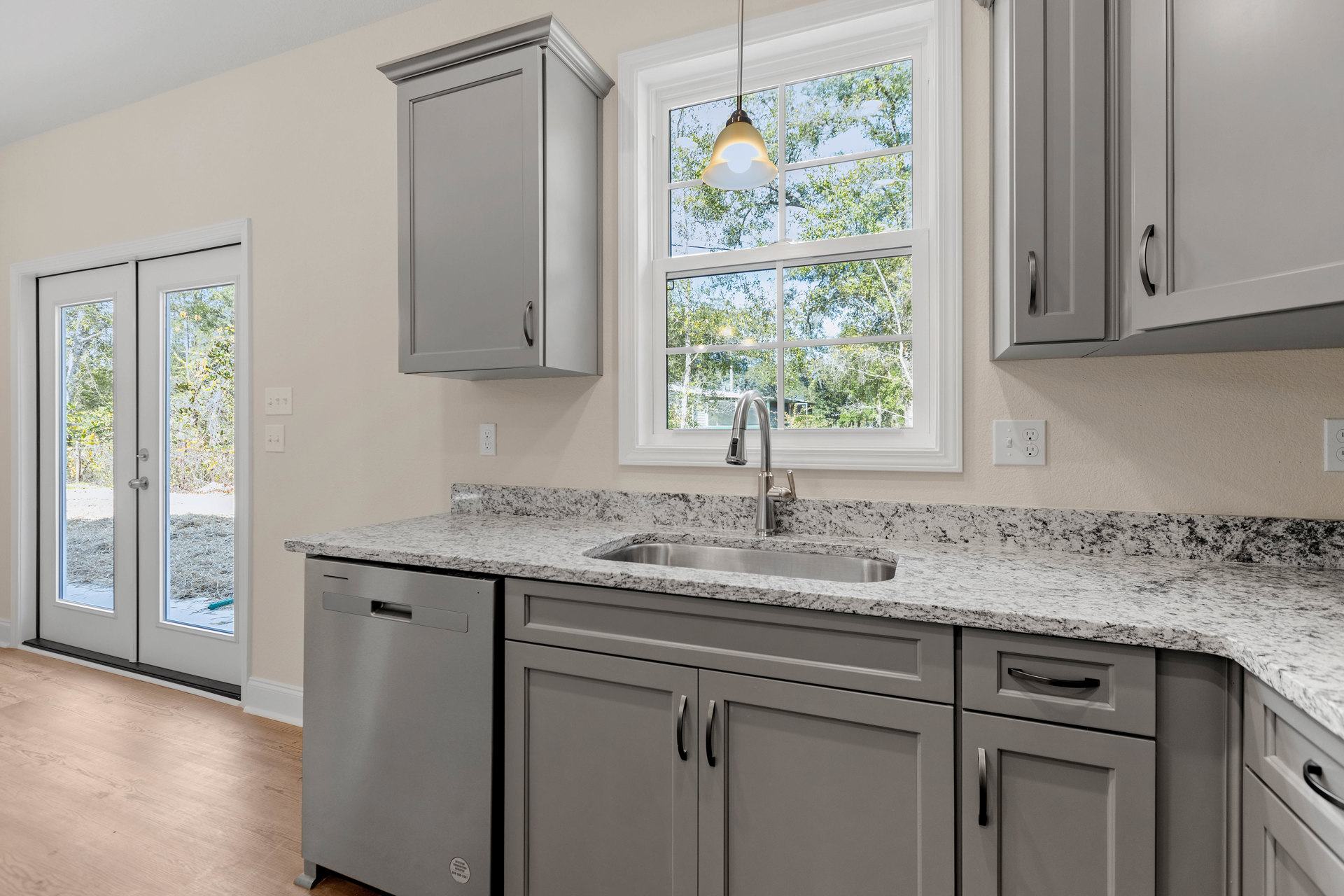 Bright kitchen featuring white cabinetry, tile backsplash, stainless steel sink beneath a window, stone countertop, glass-paned door with tree view, and ceiling light fixture.