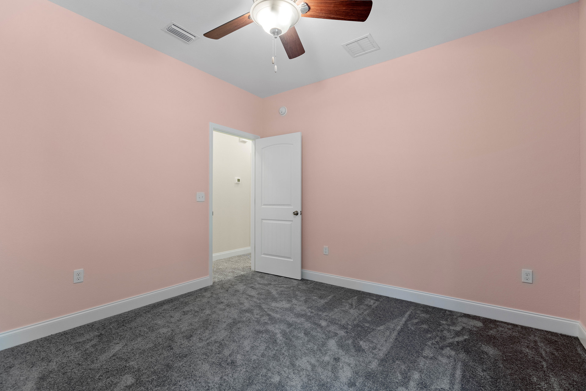 Neutral-toned room featuring a ceiling fan with light fixture, white paneled door with silver knob, smooth plaster walls, vent on the ceiling, and light-colored flooring.