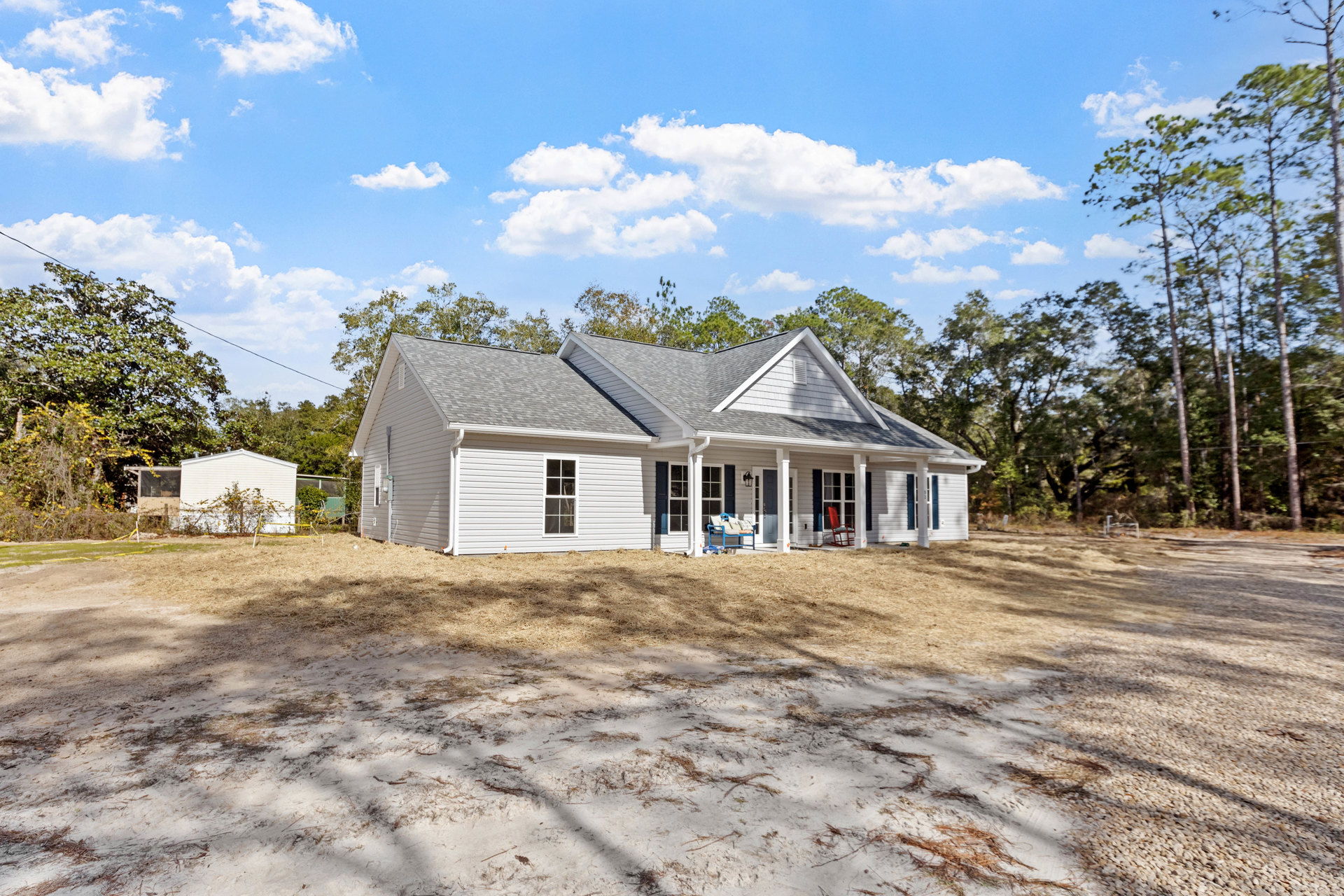 White house with blue front door, blue bench with pillows, lawn chair and swing on dirt field, fenced yard, trees and cloudy sky in background