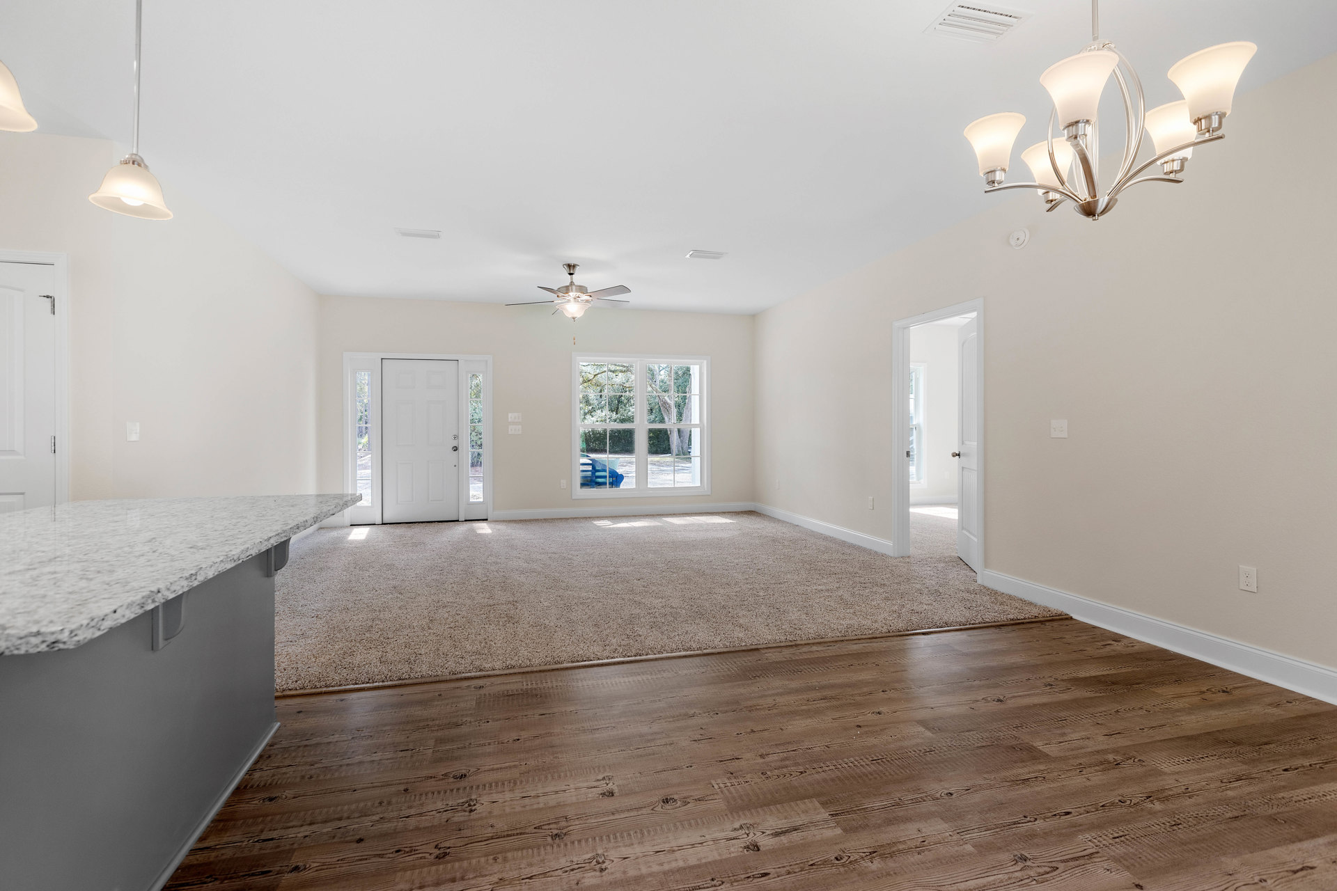 Open kitchen with marble countertop, wood flooring partially covered by a light area rug, white wall featuring exposed pipe, white door with silver handle, modern chandelier and