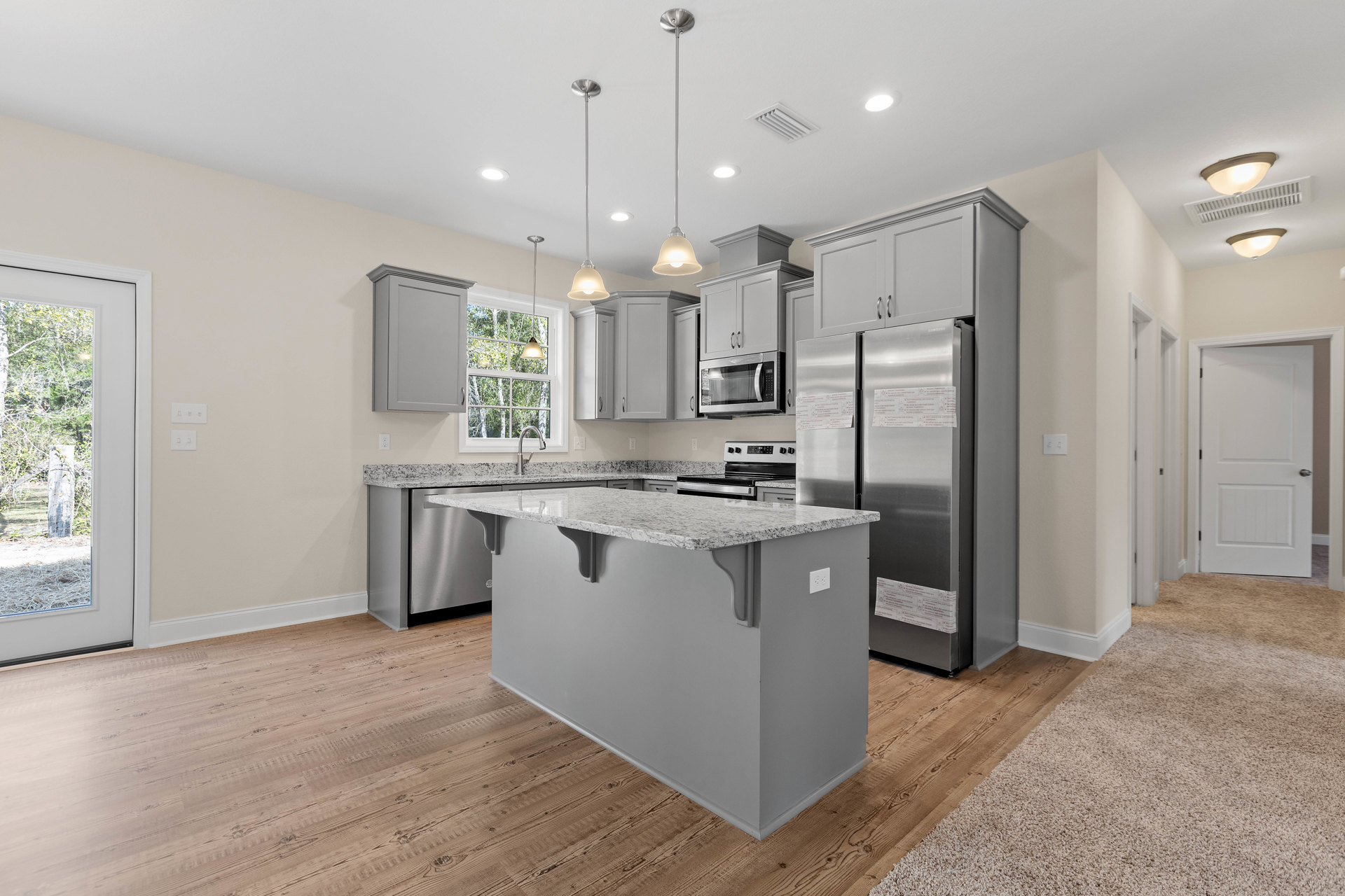 Kitchen with central island, granite countertops, stainless steel appliances, white cabinetry, microwave above stove, window overlooking fenced yard with trees, white door with