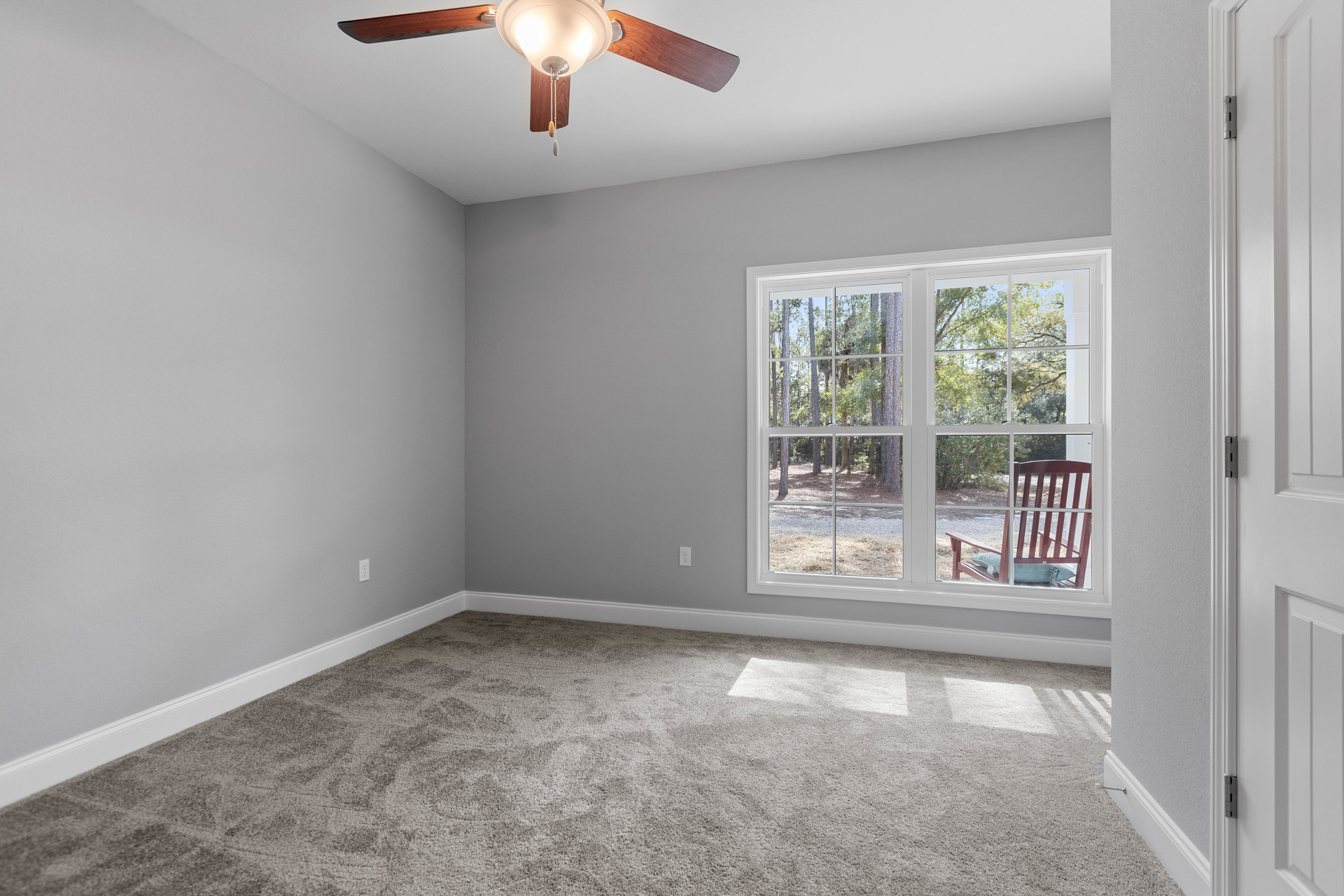 Carpeted room with white walls, ceiling fan with light fixture, large window letting in natural light, upholstered chair positioned near window