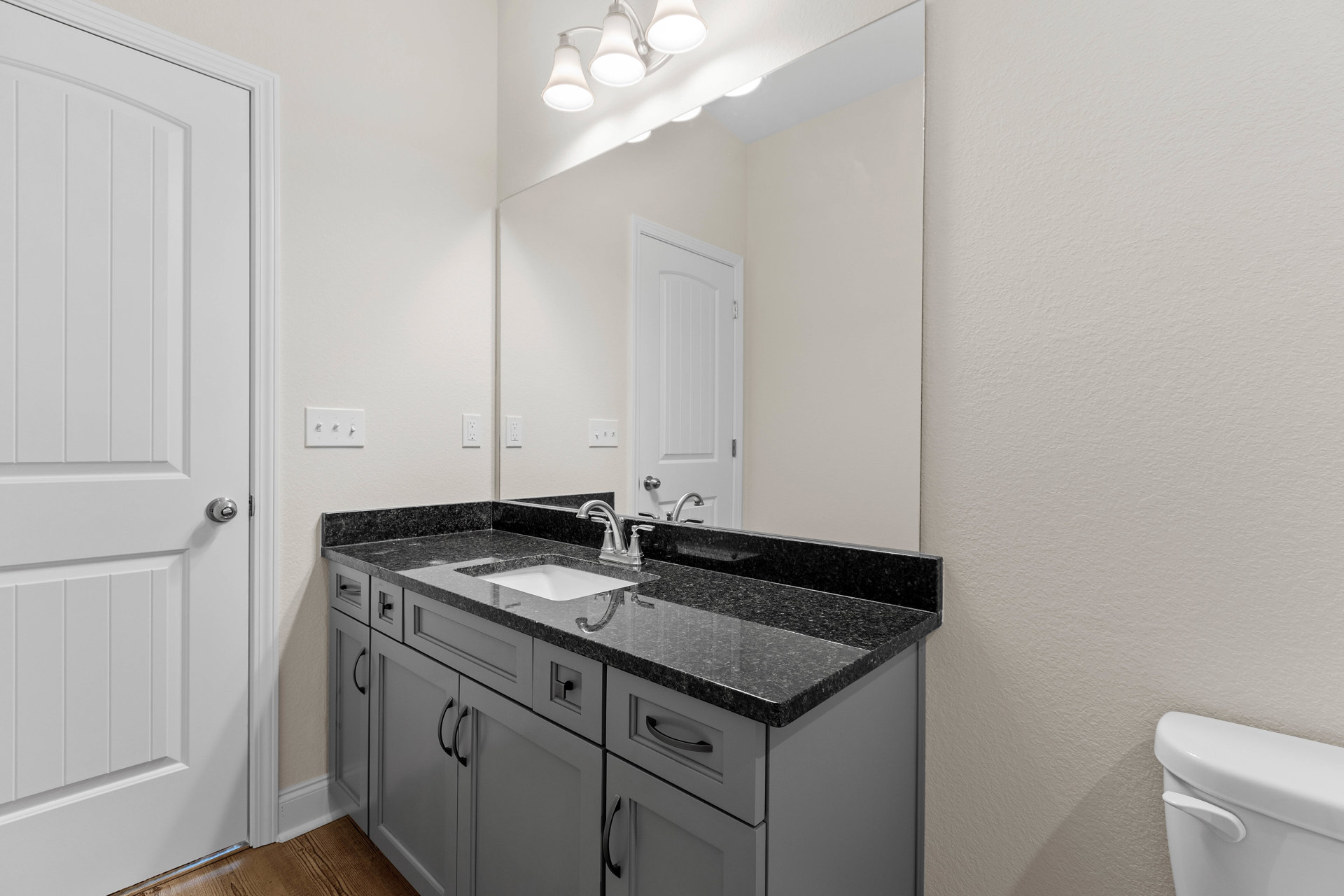 Bathroom with expansive wall mirror above white sink and wood cabinets, tiled floor, modern light fixture, white door with metal handle, and visible toilet.