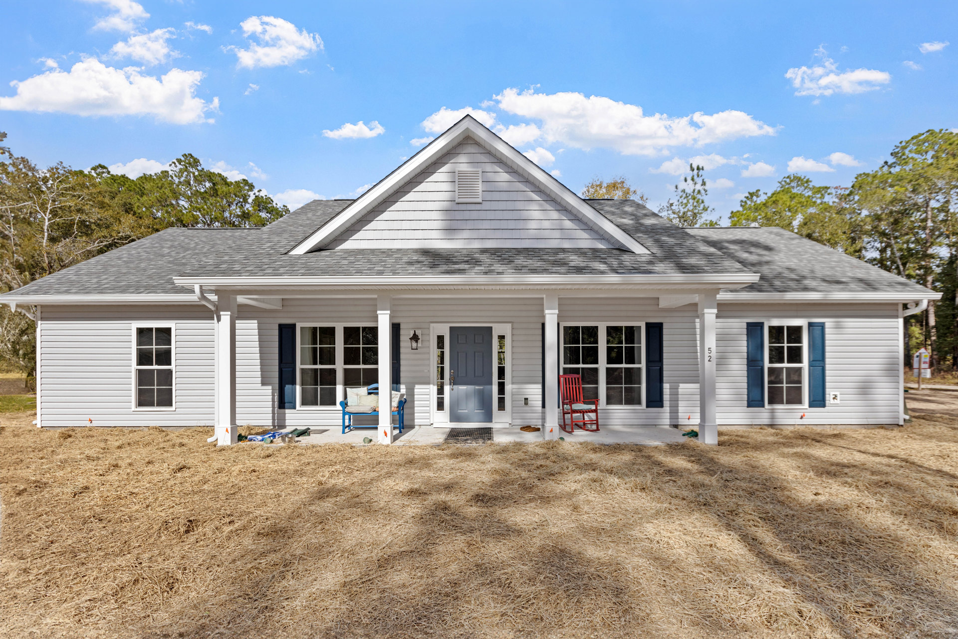 Two-story house with white siding, blue front door, grey side door, blue window shutters, white-framed windows, green lawn, red chair with pillow, blue bench with pillows, trees