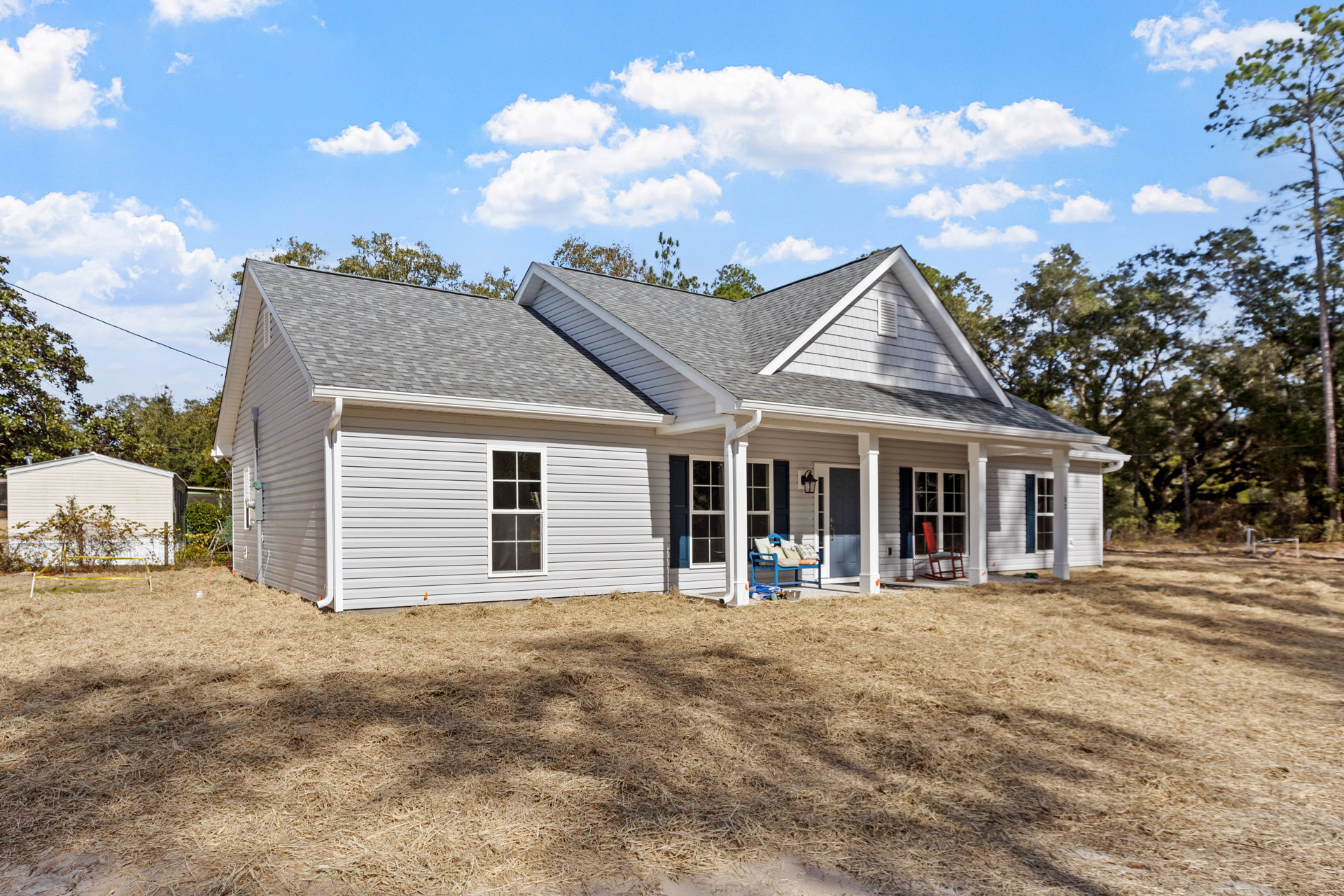 White house with blue porch, red rocking chair, and blue bench with pillows; yellow fence borders green lawn, mature trees in background