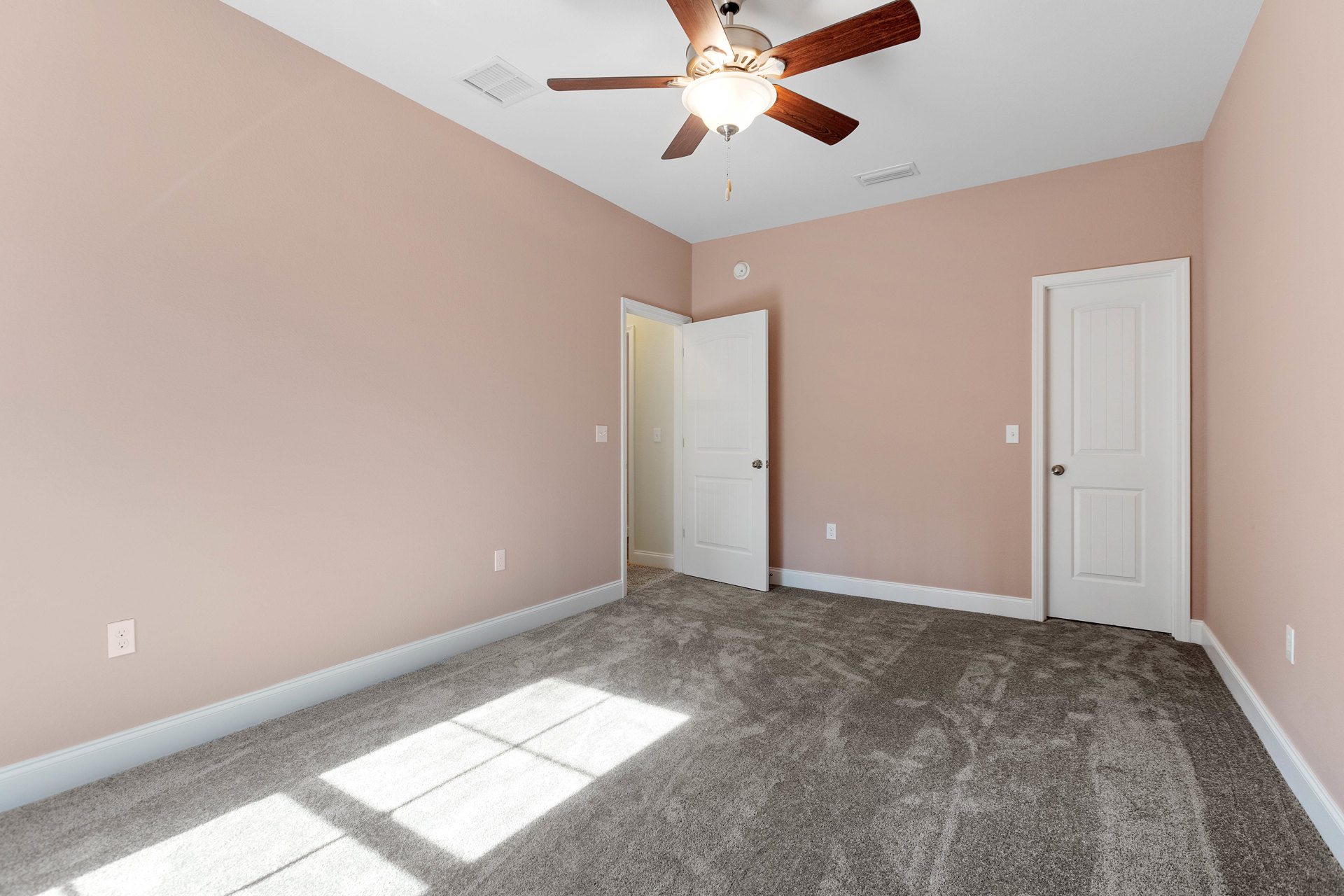 Neutral-toned carpeted room with white paneled door, silver knob, and ceiling fan featuring integrated light fixture; soft shadows cast across floor, white walls with crown