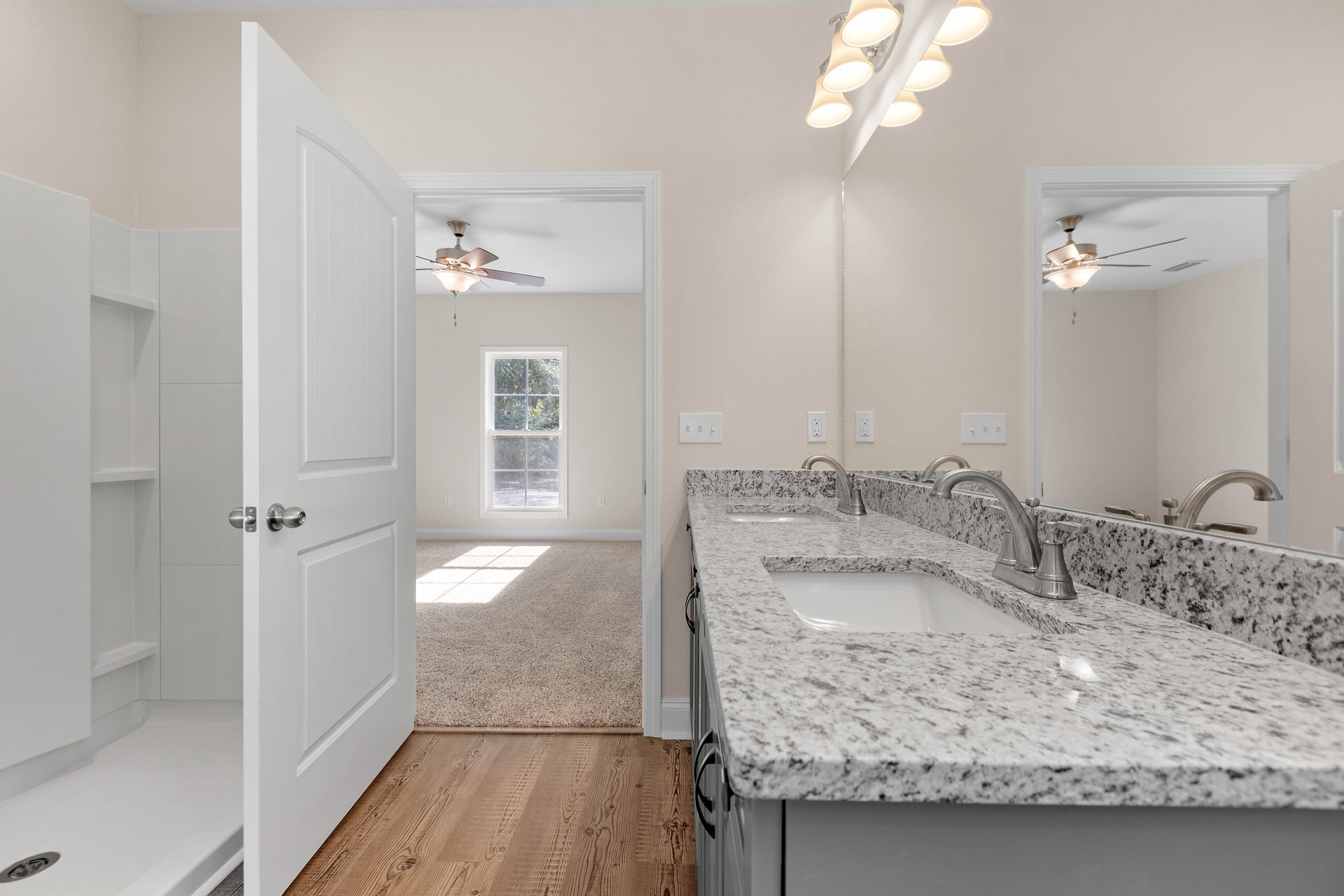 Bathroom with marble countertops, wood flooring, white sink with silver faucet, ceiling lights, large window overlooking trees, and door with silver knob