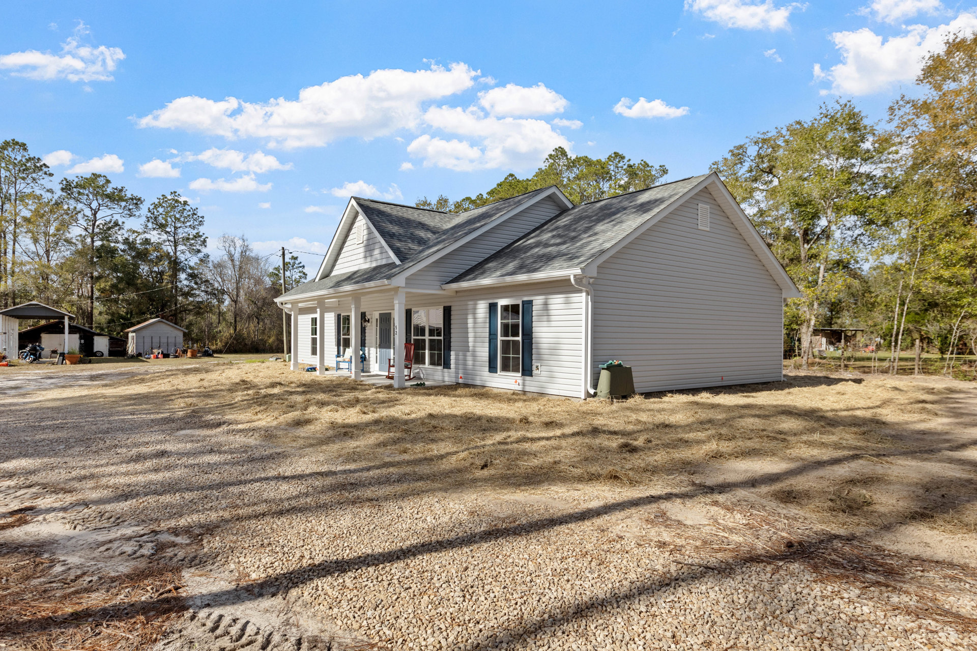 White siding house with blue shutters, large covered porch, white-framed windows, green wheeled trash can near entry, shingled roof, mature trees in background.