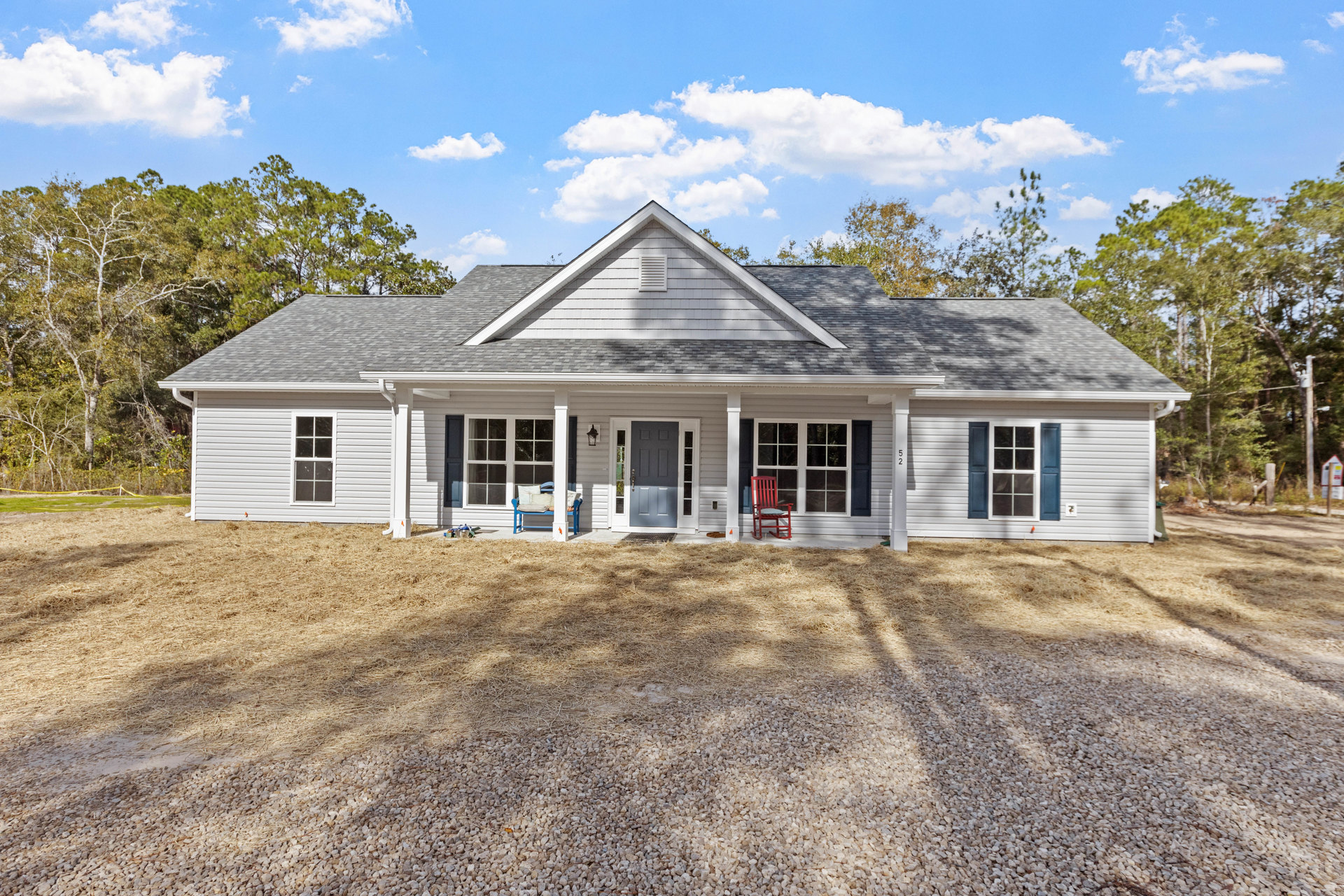 Gravel driveway leading to a cottage-style home with blue and gray doors, red rocking chair and blue bench with pillows on the porch, surrounded by trees under a cloudy sky