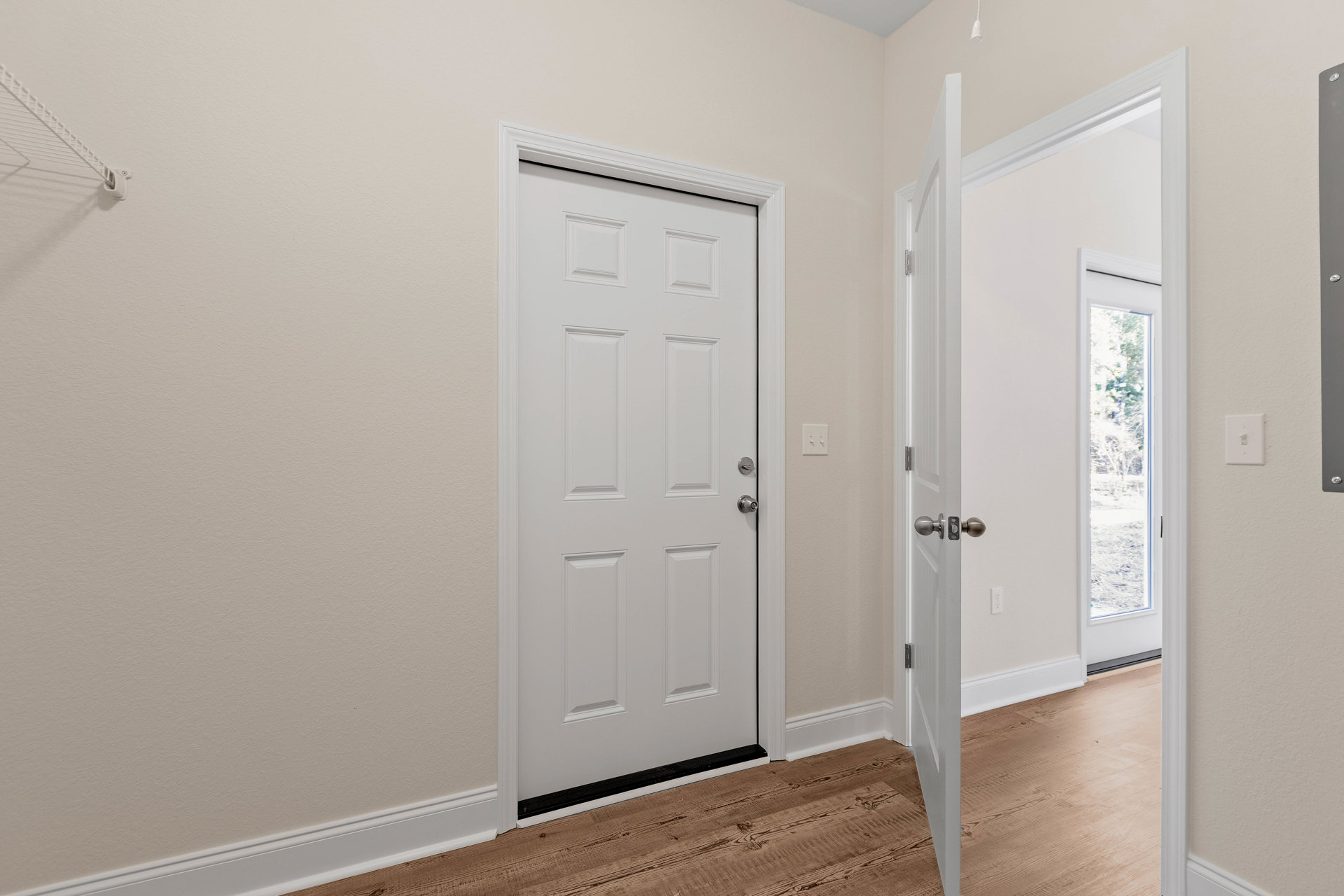 White door with silver knobs and black trim, light switch on adjacent wall, window revealing tree outside, hardwood flooring and white ceiling in residential interior.
