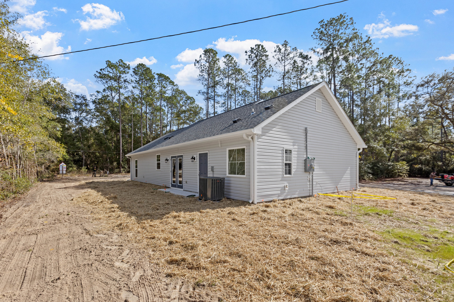 White house with large windows reflecting surrounding trees, black heat pump unit on dry grass yard, mature trees in background under cloudy sky