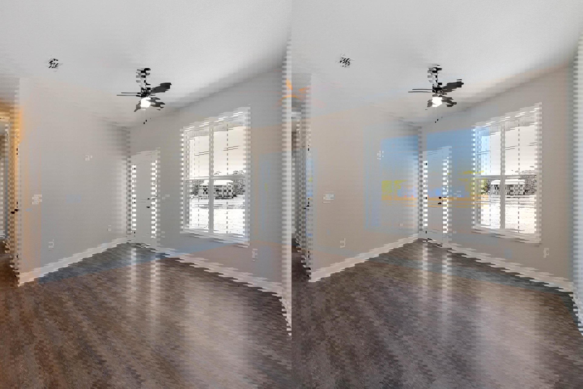 Room with wood flooring, white walls, ceiling fan with light, large window showing neighboring house, and white door with black hardware
