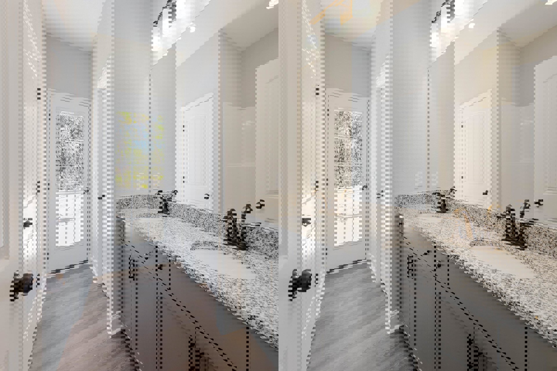 Bathroom with double sinks on granite countertop, wood flooring, white cabinetry, and a door opening to a view of trees.