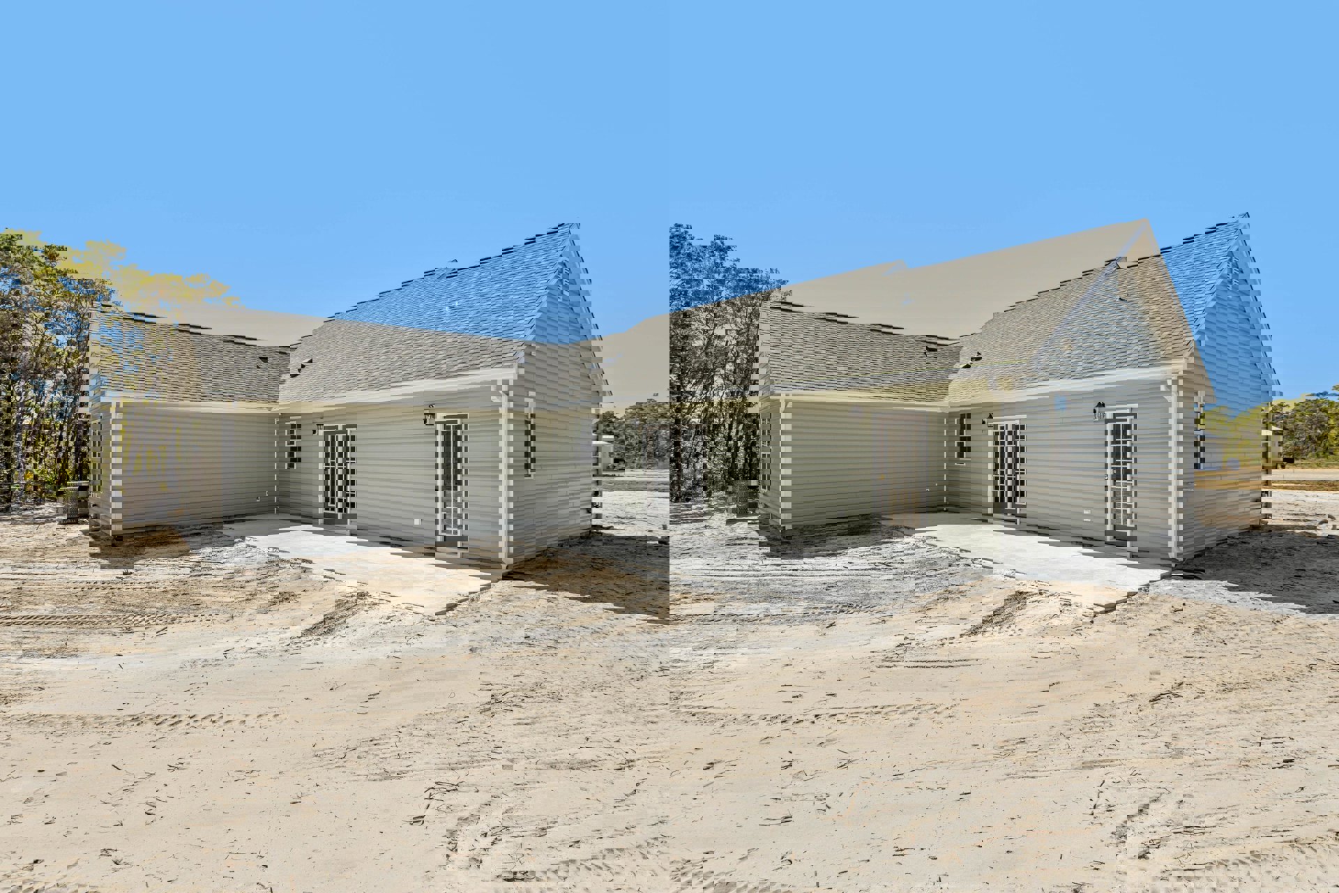 Two-story home with light siding, glass-paneled front door, paved driveway showing tire tracks, small patio area, and mature trees in the yard