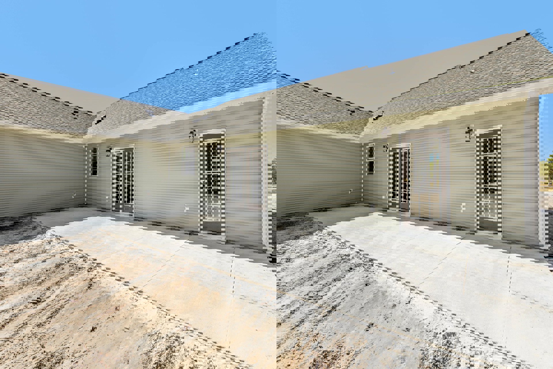 Modern two-story home with light-colored siding, prominent front door, large windows, concrete driveway bordered by dirt and sand, shingled roof, and blue sky overhead