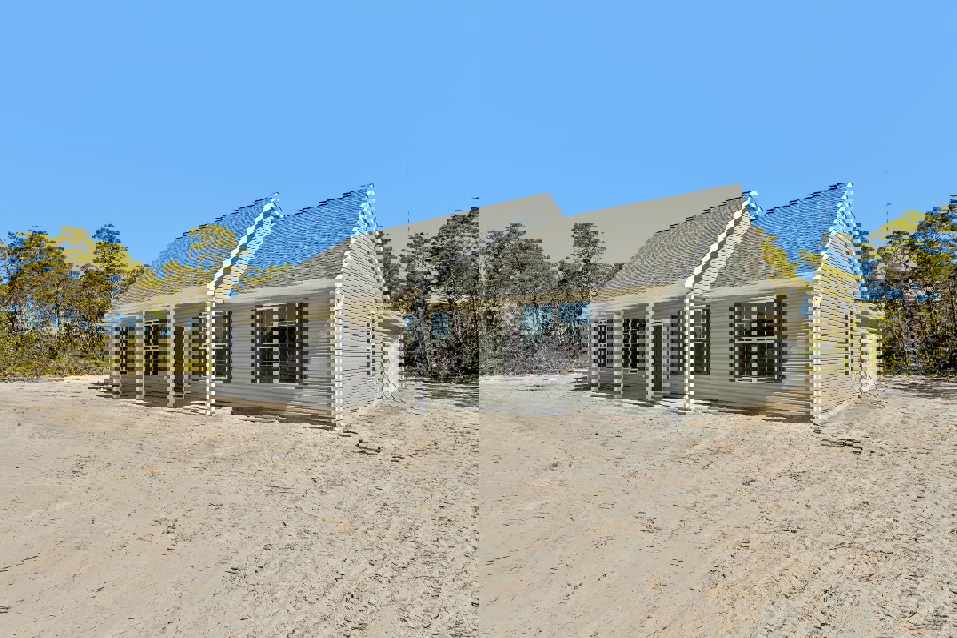 Two-story white house with white columns and porch, white-framed windows, dirt yard in front, blue sky overhead, and trees in the background