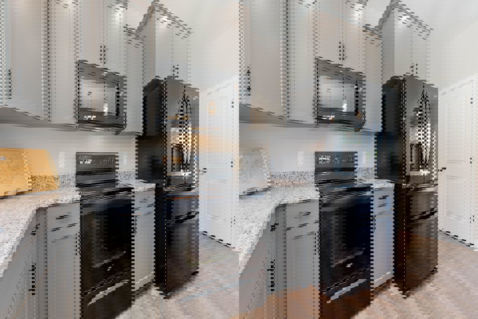 Grey kitchen cabinets with black double-door refrigerator, black stove with glass door, black microwave under ceiling light, wood cutting board with engraved name on countertop