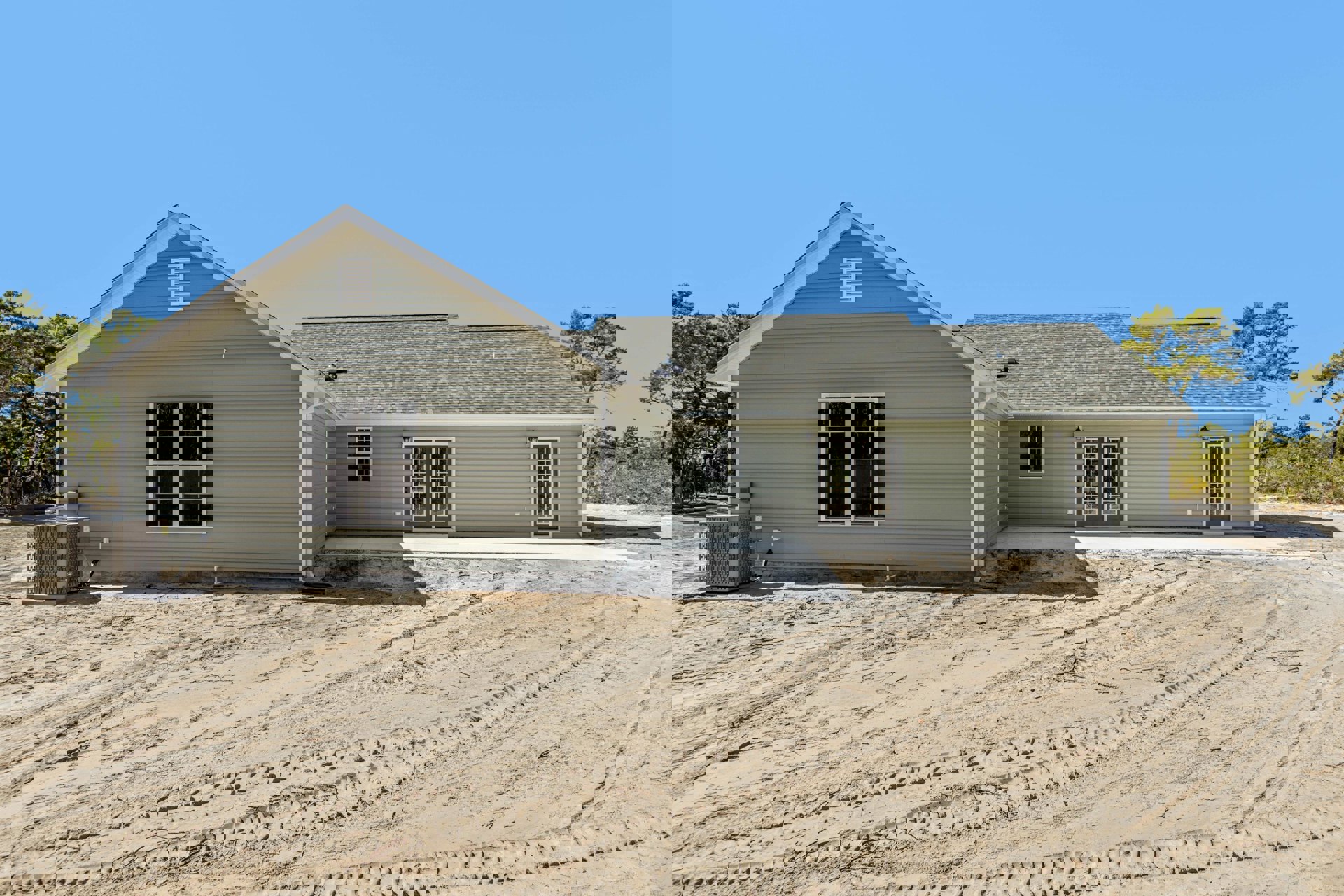 Two-story home with light siding, multiple-paned windows, white-framed double doors, dirt yard with tire tracks, and sparse landscaping under a clear sky