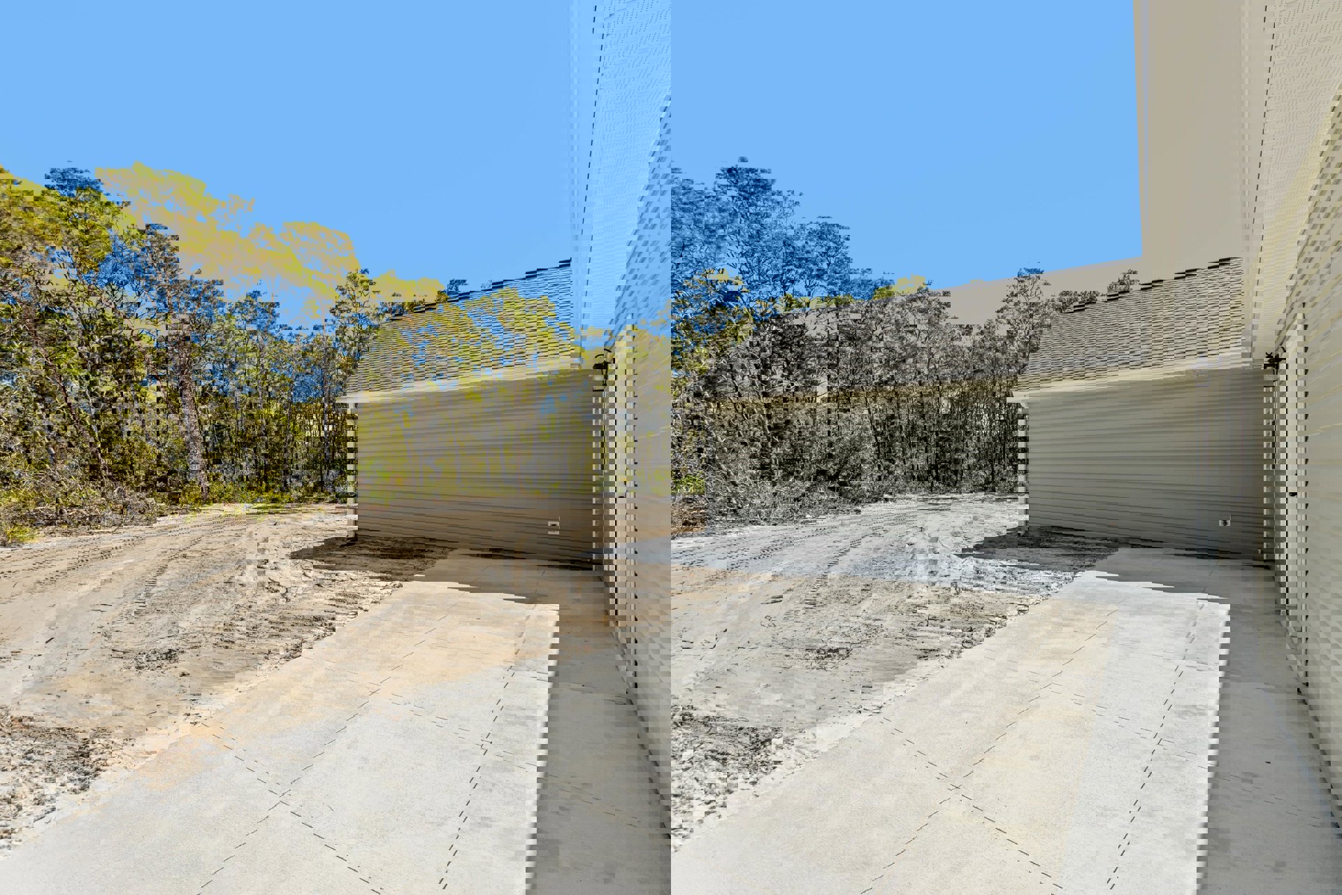 Concrete driveway leading to a garage, surrounded by mature trees and blue sky, with composite roof and dirt landscaping visible