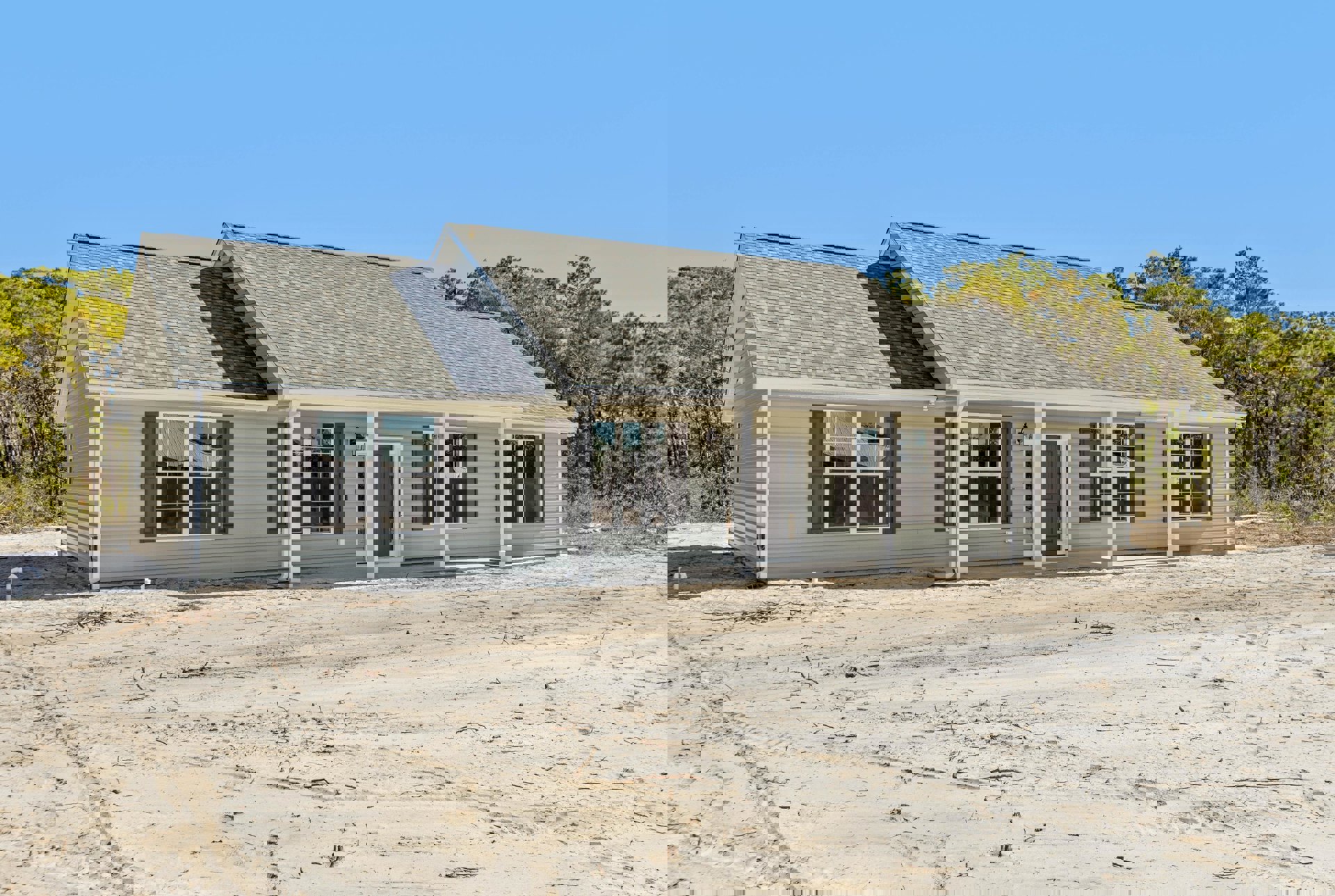 White-roofed house with attached garage, covered porch featuring a light fixture, dirt driveway and yard, large windows, surrounded by trees under a clear sky