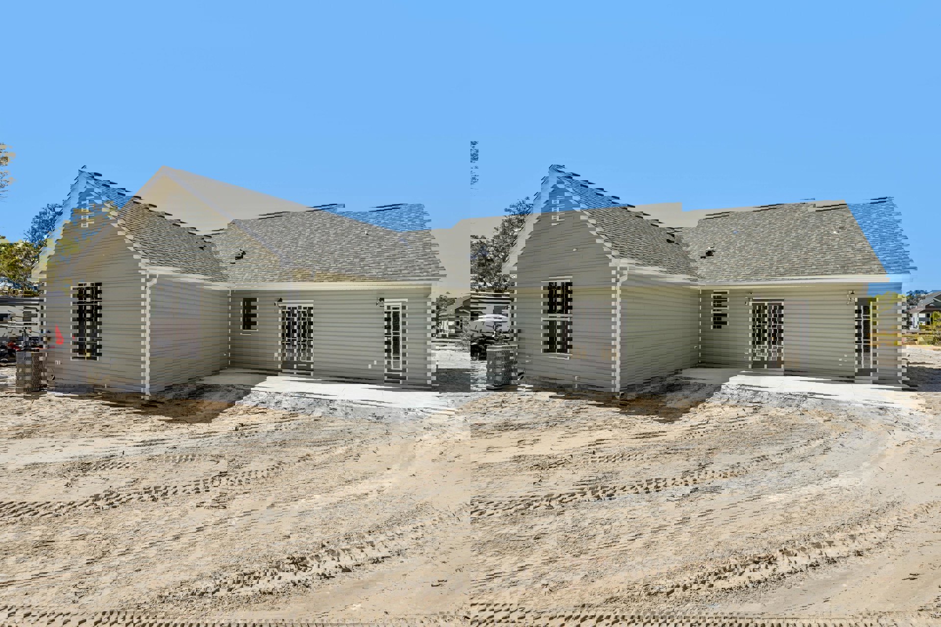 Modern home exterior with light siding, double front doors featuring glass panels, large windows with multiple panes, dirt yard with visible tire tracks, and surrounding trees