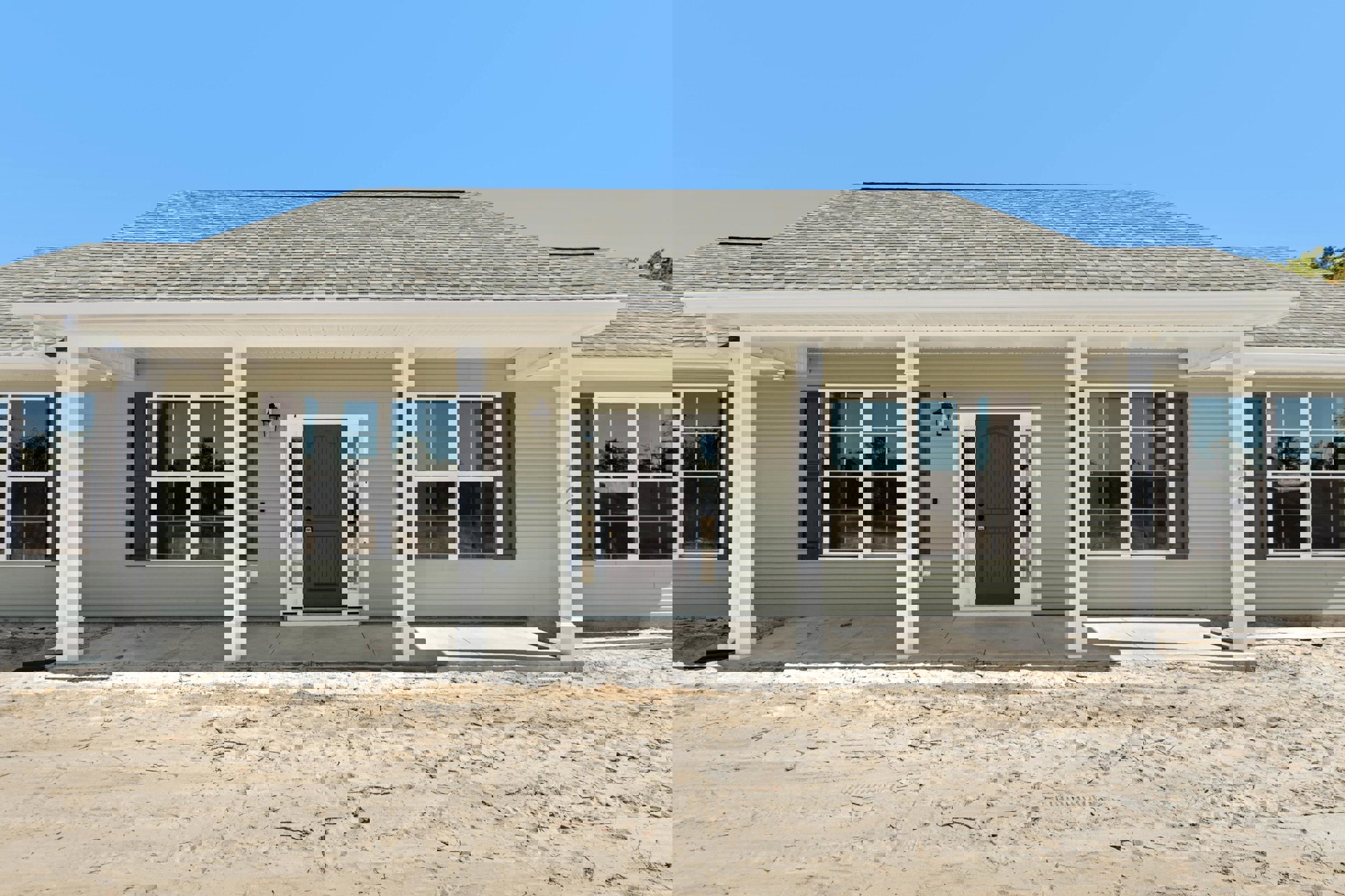 Front view of a house with light siding, grey door framed in white, glass-paned windows, and a sandy dirt road leading to the garage door