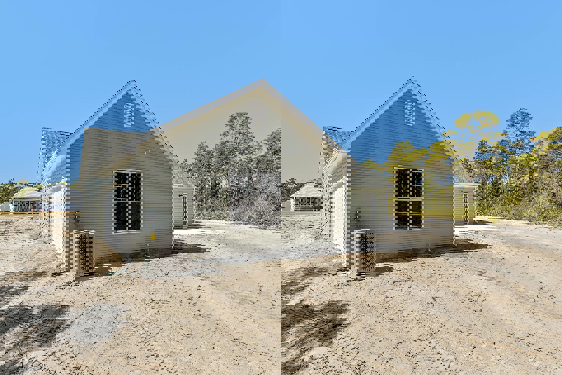 Two-story house with attached garage, multi-pane windows, large gray heat pump, white exterior vent, pitched roof, dirt driveway, and surrounding trees.