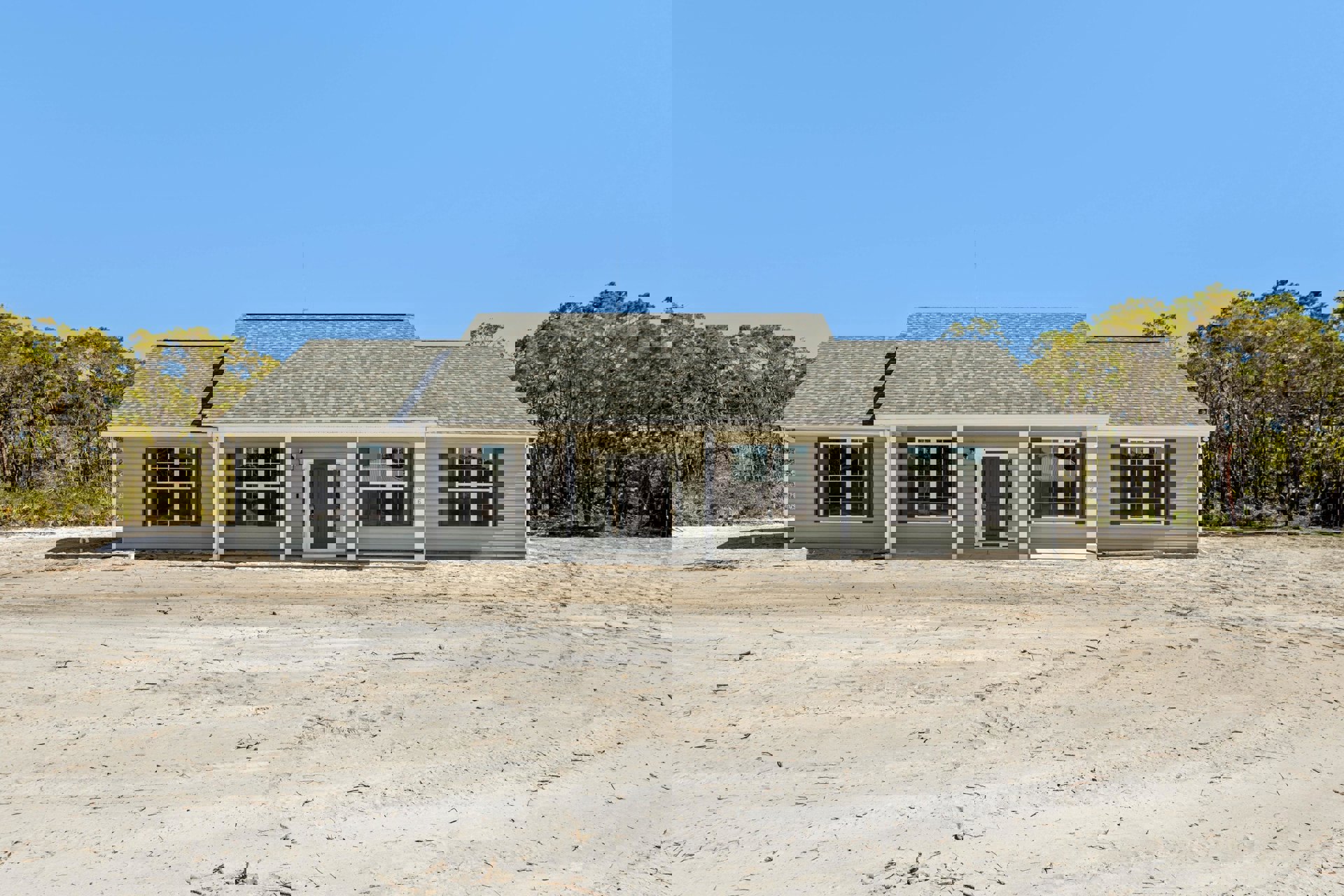 Single-story house with white roof, grey door, and multiple windows, surrounded by trees and set on a dirt lot; antenna visible atop the roof.